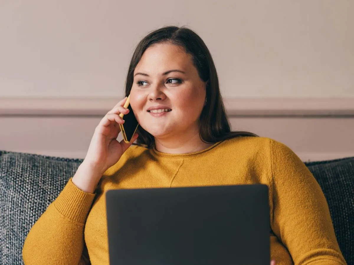 Frau in gelbem Pullover sitzt mit Laptop auf dem Schoß und telefoniert lächelnd.