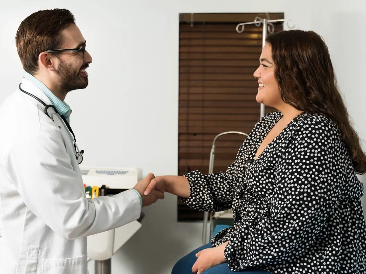 Arzt mit weißem Kittel und Stethoskop schüttelt lächelnd die Hand einer sitzenden Patientin.