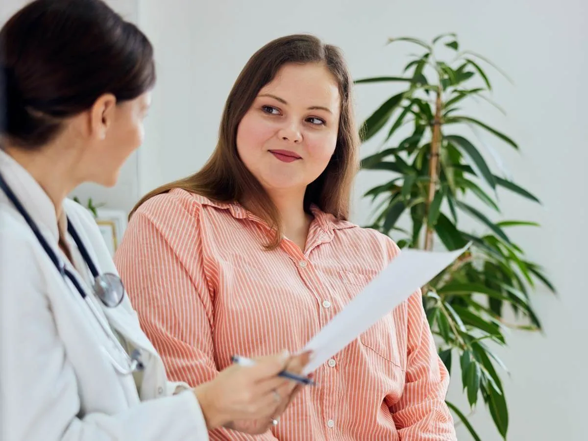 Eine Ärztin im weißen Kittel bespricht mit einer Frau in rosa gestreiftem Hemd ein Dokument im hellen Behandlungszimmer.