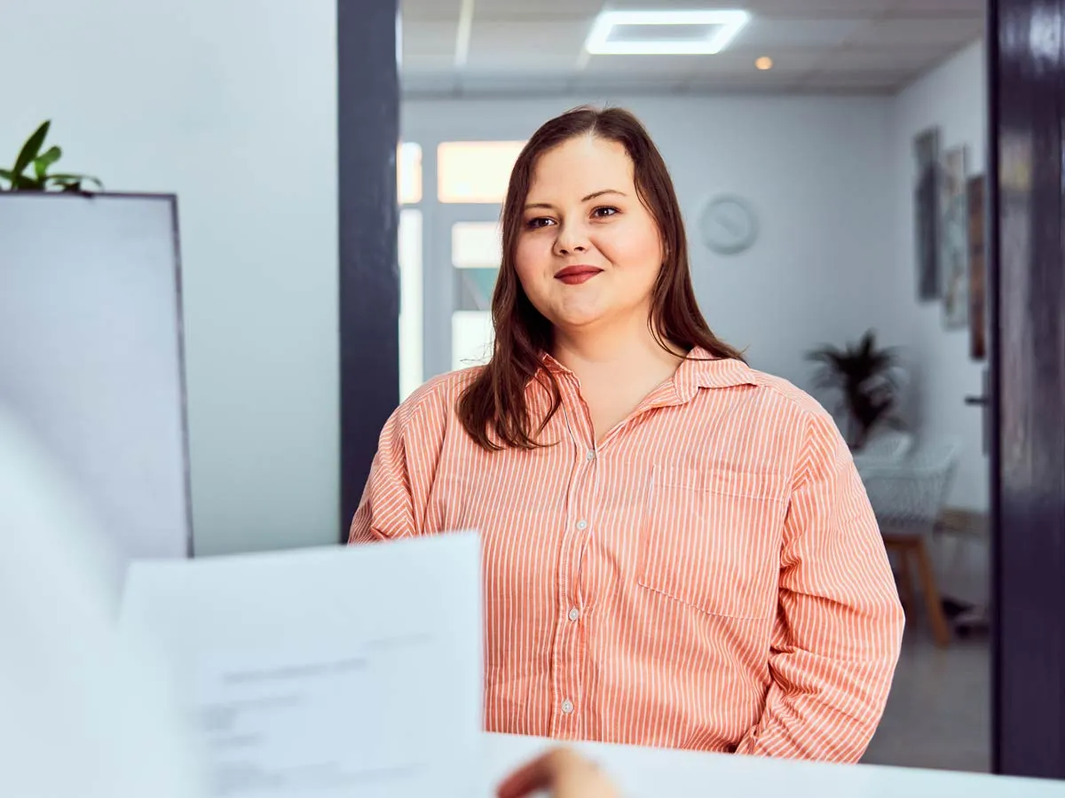 Junge Frau mit orange-weiß gestreiftem Hemd steht lächelnd an einem Tresen in einem modernen Büro.