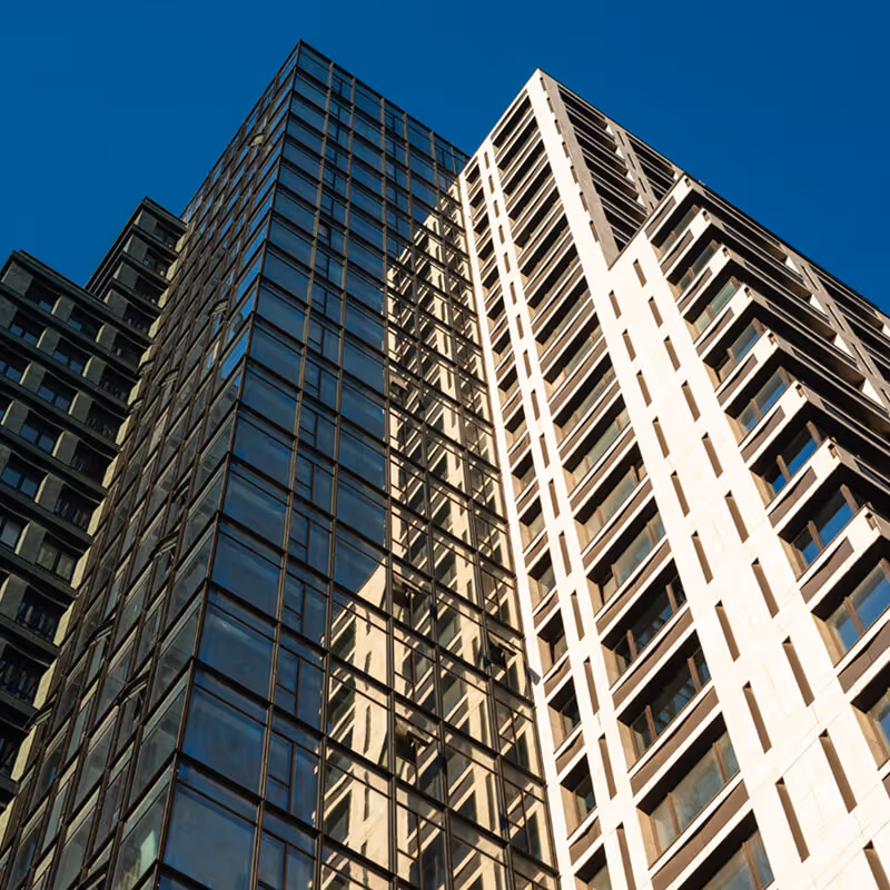 A man in a white dress shirt and tie sits in a high-rise office. Behind him, a large window with blinds reveals a prominent view of the New York City skyline and the Empire State Building in golden sunlight.