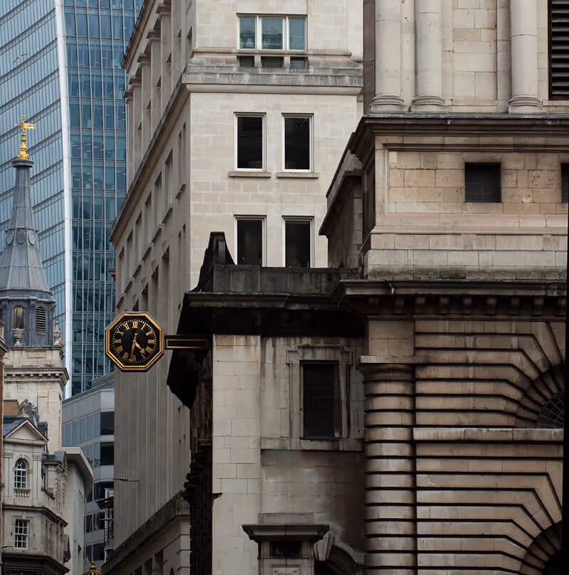 A detailed architectural shot in a financial district, showing the contrast between a classic stone building with a prominent gold-rimmed outdoor clock and a soaring, modern glass-and-steel skyscraper in the background.