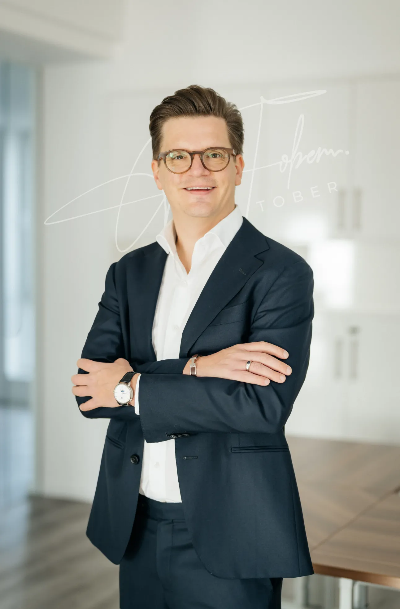 Porträt eines Mannes mit Brille im dunklen Anzug und weißem Hemd, der mit verschränkten Armen lächelt vor einem modernen Bürohintergrund.