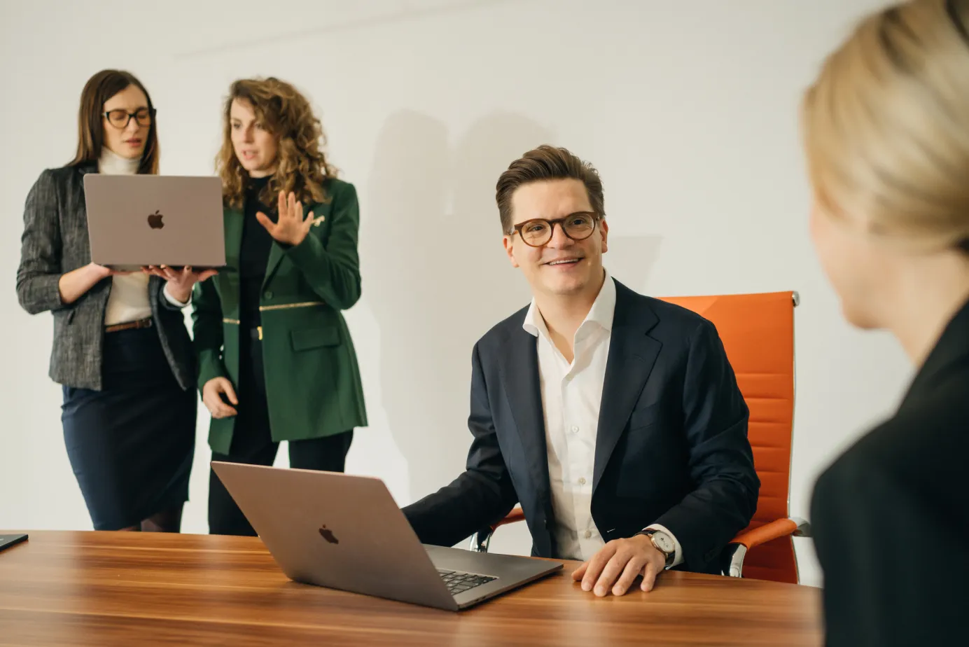 Ein Mann in Brille und Anzug sitzt an einem Tisch mit Laptop und lächelt eine Frau gegenüber an, während zwei Frauen im Hintergrund einen Laptop anschauen und diskutieren.