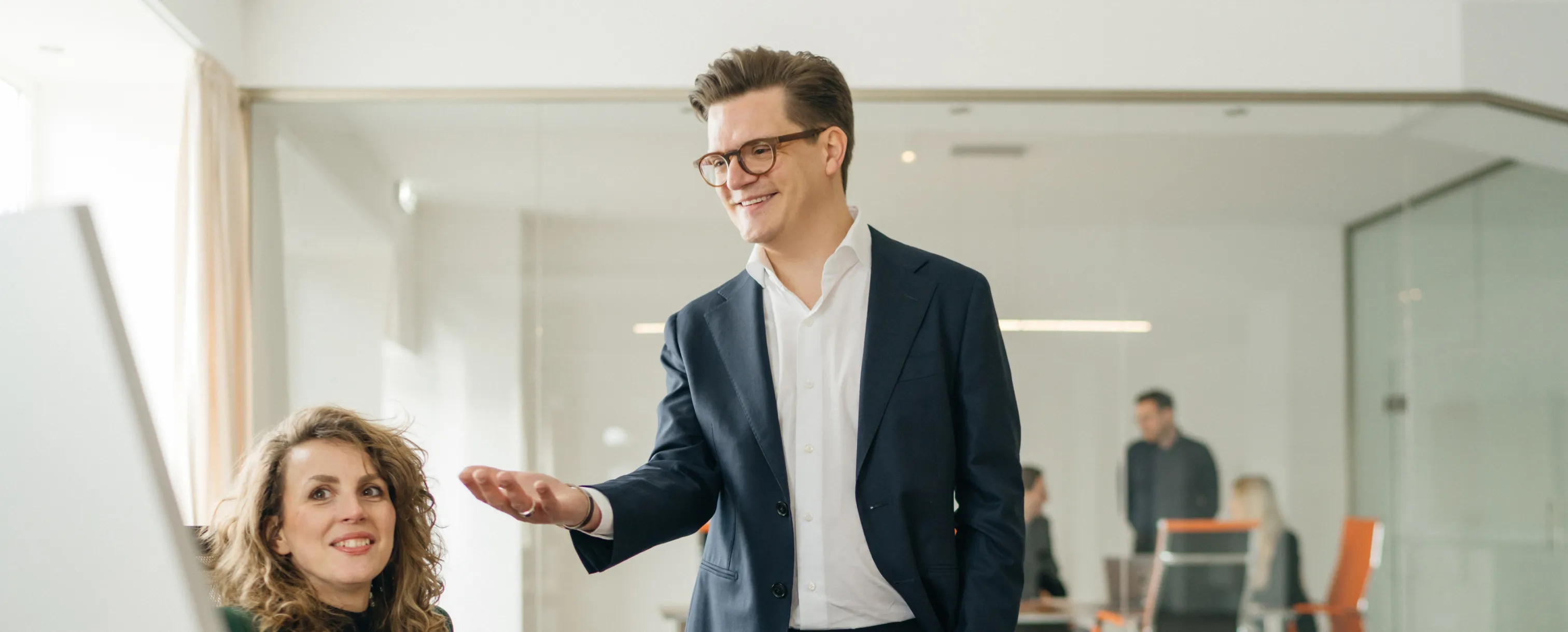 Ein lächelnder Mann im Anzug erklärt einer Frau mit lockigen Haaren am Schreibtisch etwas in einem modernen Büro.