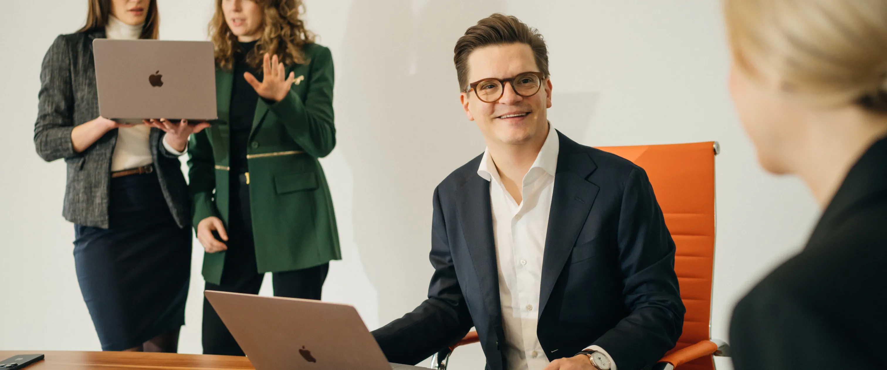 Ein lächelnder Mann in Anzug und Brille sitzt am Tisch mit einem Laptop, während drei Frauen mit Laptop und Gespräch im Hintergrund stehen.