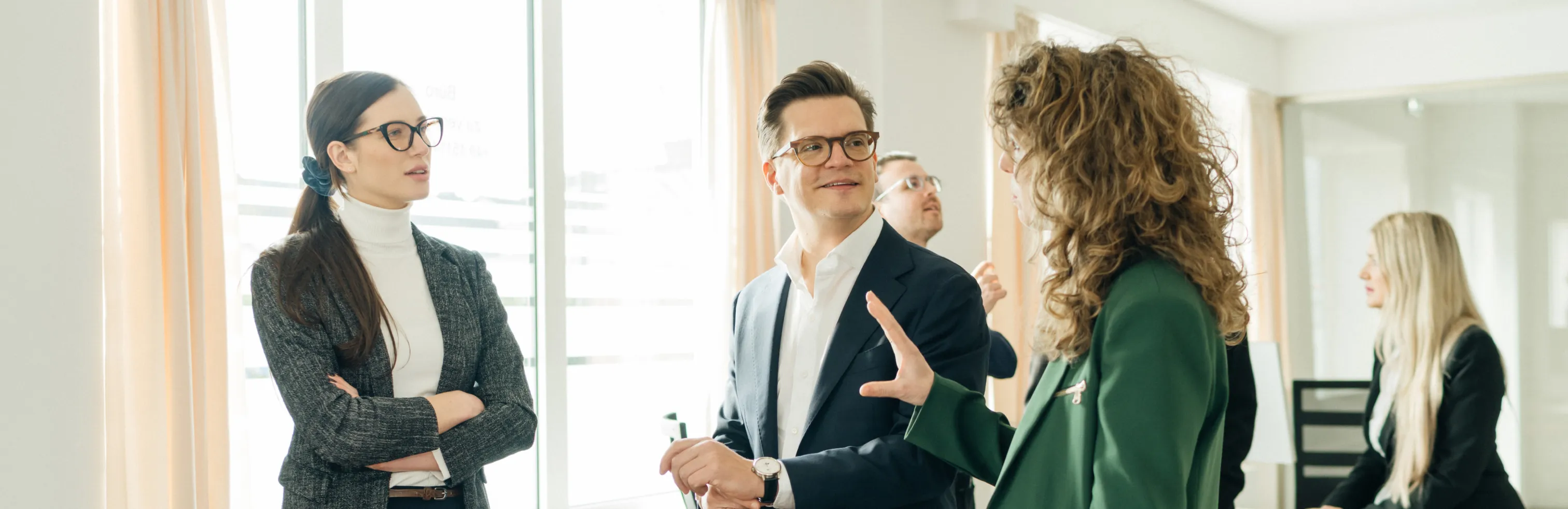 Fünf moderne Berufstätige diskutieren in einem hellen Büro, zwei Frauen und ein Mann sprechen im Vordergrund, zwei weitere Personen im Hintergrund.