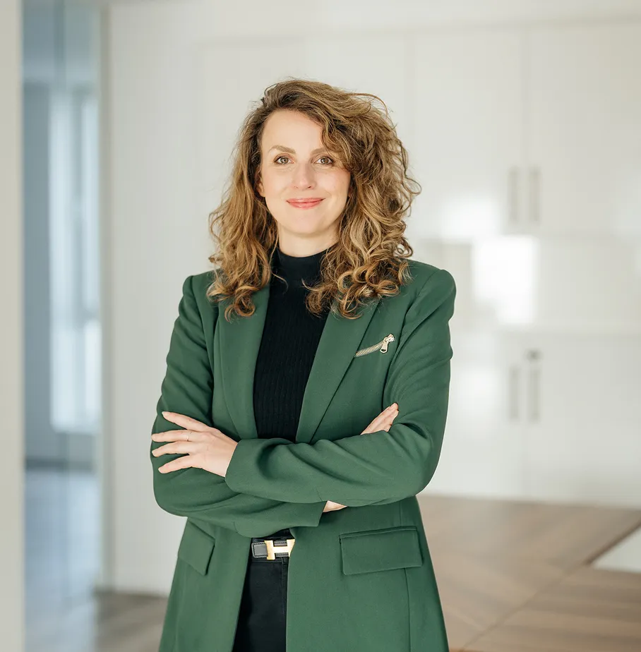 Frau mit lockigem braunem Haar, die einen dunkelgrünen Blazer und einen schwarzen Pullover trägt, mit verschränkten Armen vor weißem Hintergrund.