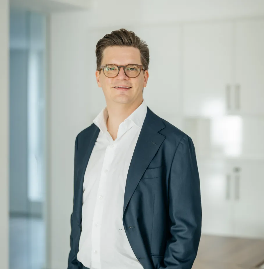 Frau mit lockigem braunem Haar, die einen dunkelgrünen Blazer und einen schwarzen Pullover trägt, mit verschränkten Armen vor weißem Hintergrund.