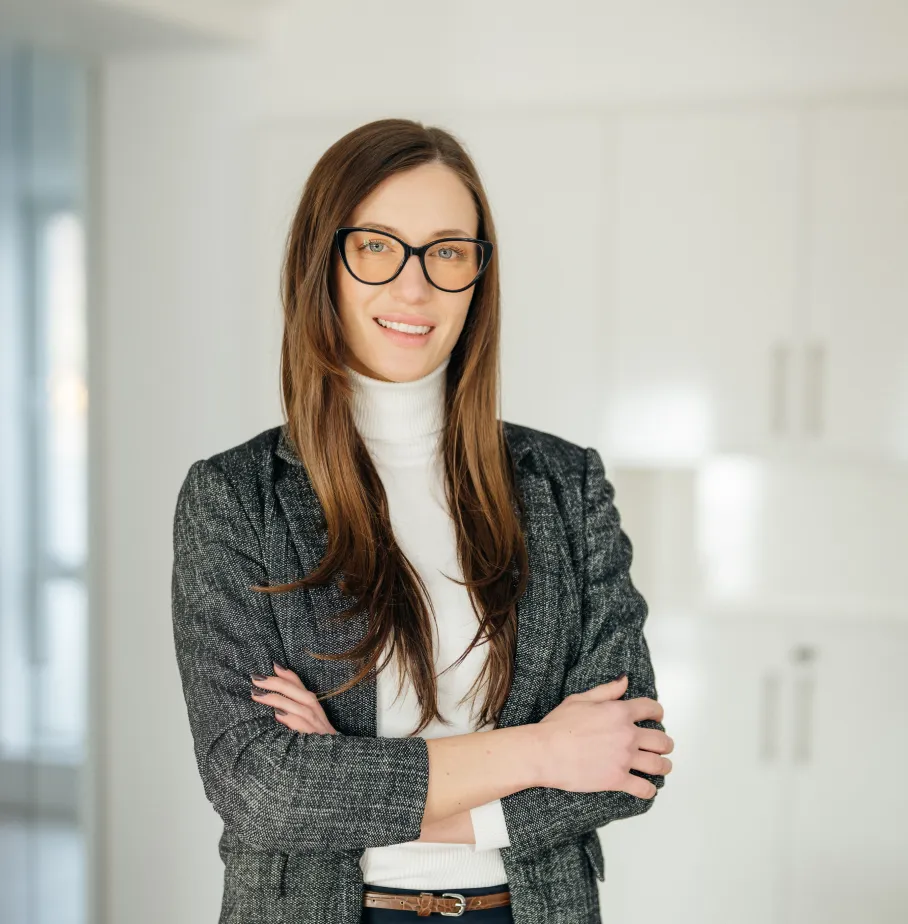 Frau mit lockigem braunem Haar, die einen dunkelgrünen Blazer und einen schwarzen Pullover trägt, mit verschränkten Armen vor weißem Hintergrund.
