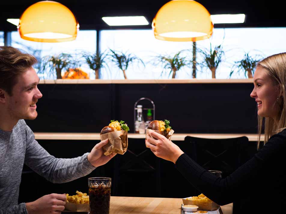 Nuori mies ja nainen hymyilevät ja kilistävät hampurilaisilla ravintolapöydässä, jonka ääressä on ruokaa ja juomia.