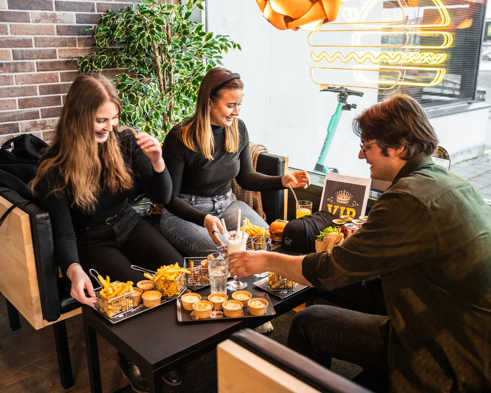 Kolme ystävää istuu kahvilapöydän ympärillä ranskalaisten, juomien ja kastikkeiden kanssa, nauttien ateriastaan yhdessä.
