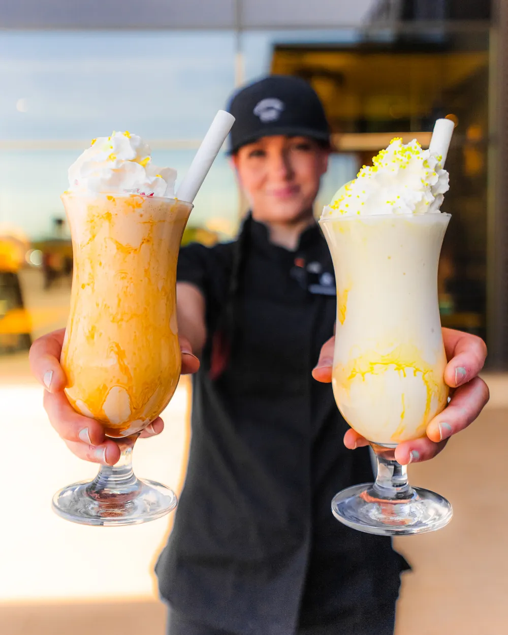 Henkilö mustissa pitää kahta kermavaahdolla kuorrutettua pirtelöä, joissa on keltaisia kastikkeita ja pillit, kohti kameraa.