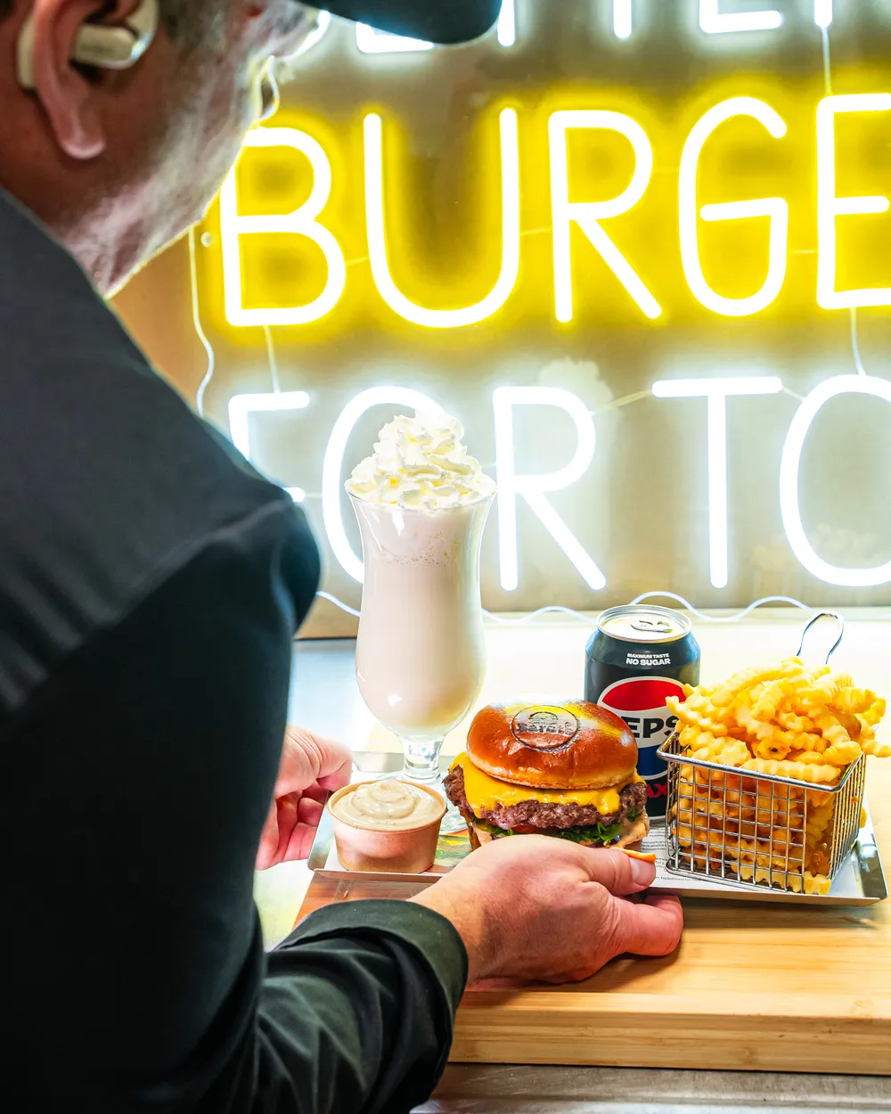 Henkilö pitelee tarjotinta, jolla on juustohampurilainen, uritetut ranskalaiset metallikorissa, tölkki Pepsi Maxia, milkshake kermavaahdolla ja pieni kuppi kastiketta.