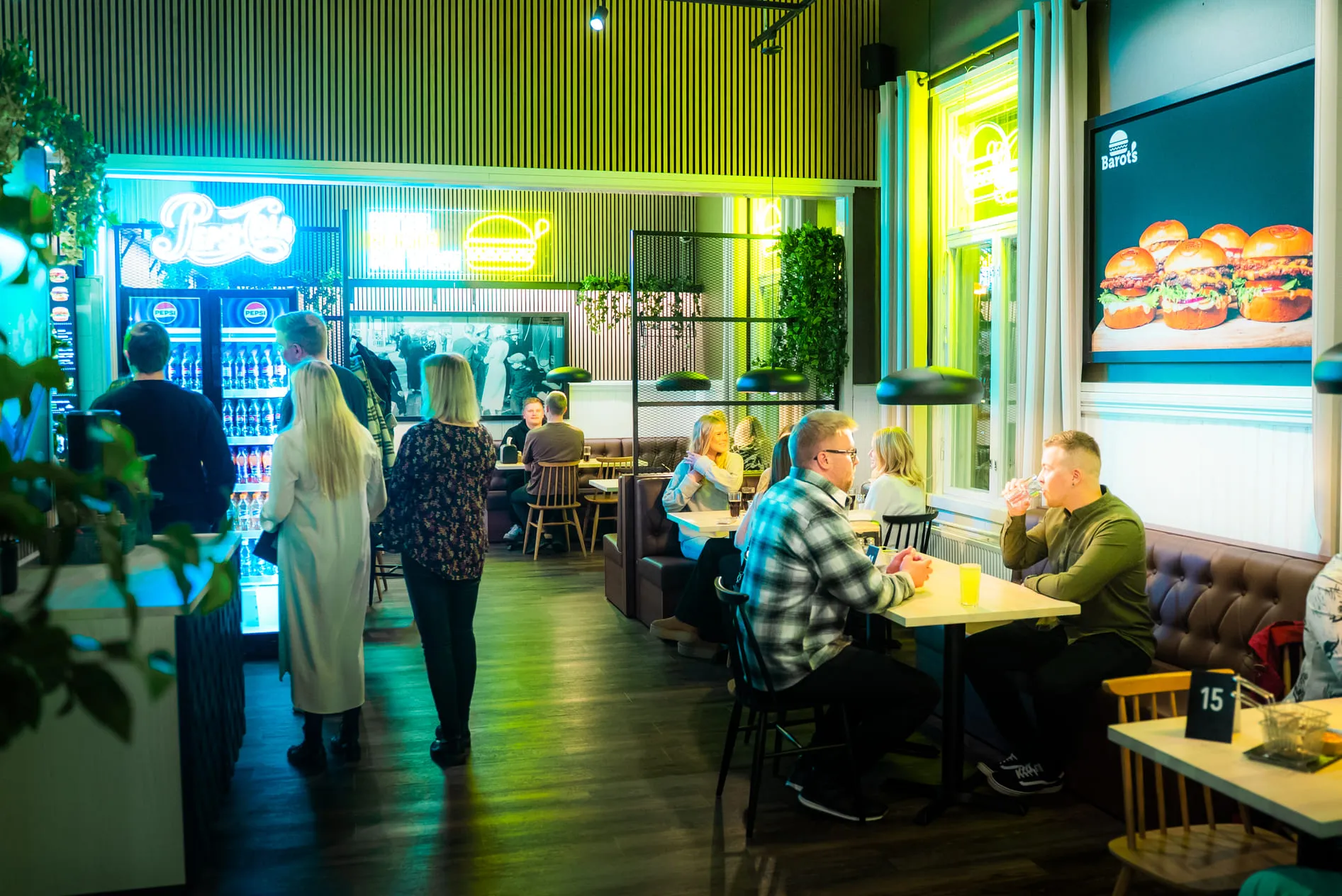 Ihmisiä syömässä ja seisomassa modernissa ravintolassa, jossa on neonkylttejä ja burger-mainos seinällä.