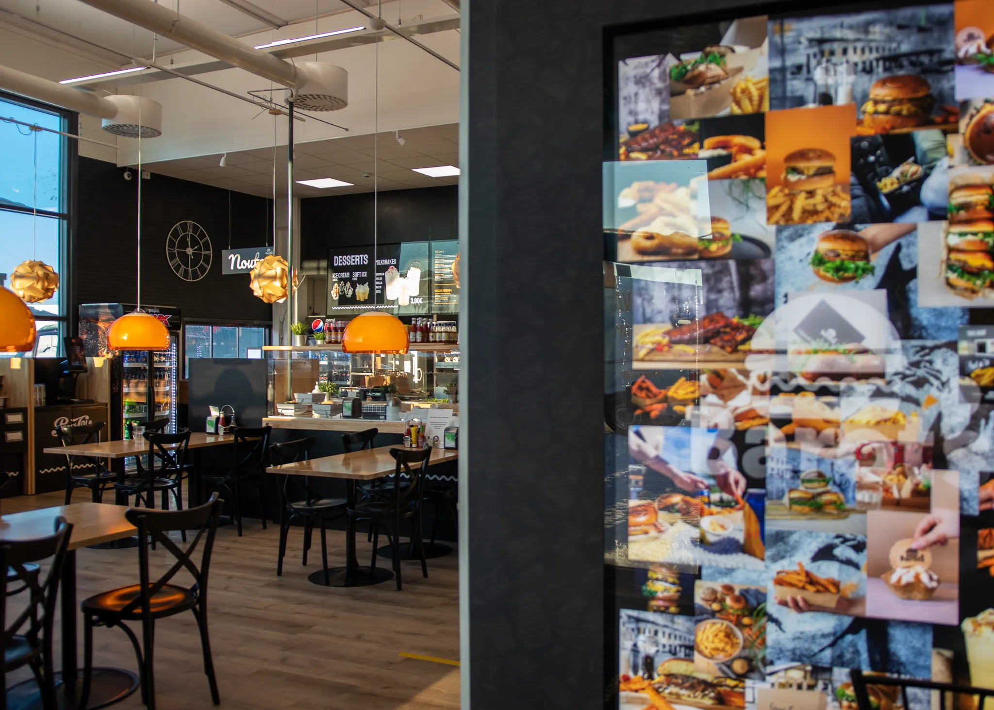 Sisäkuva modernista pikaruokaravintolasta, jossa on puisia pöytiä, mustia tuoleja, oransseja riippuvalaisimia ja digitaalimenu jälkiruokien esittelyyn tiskin takana.