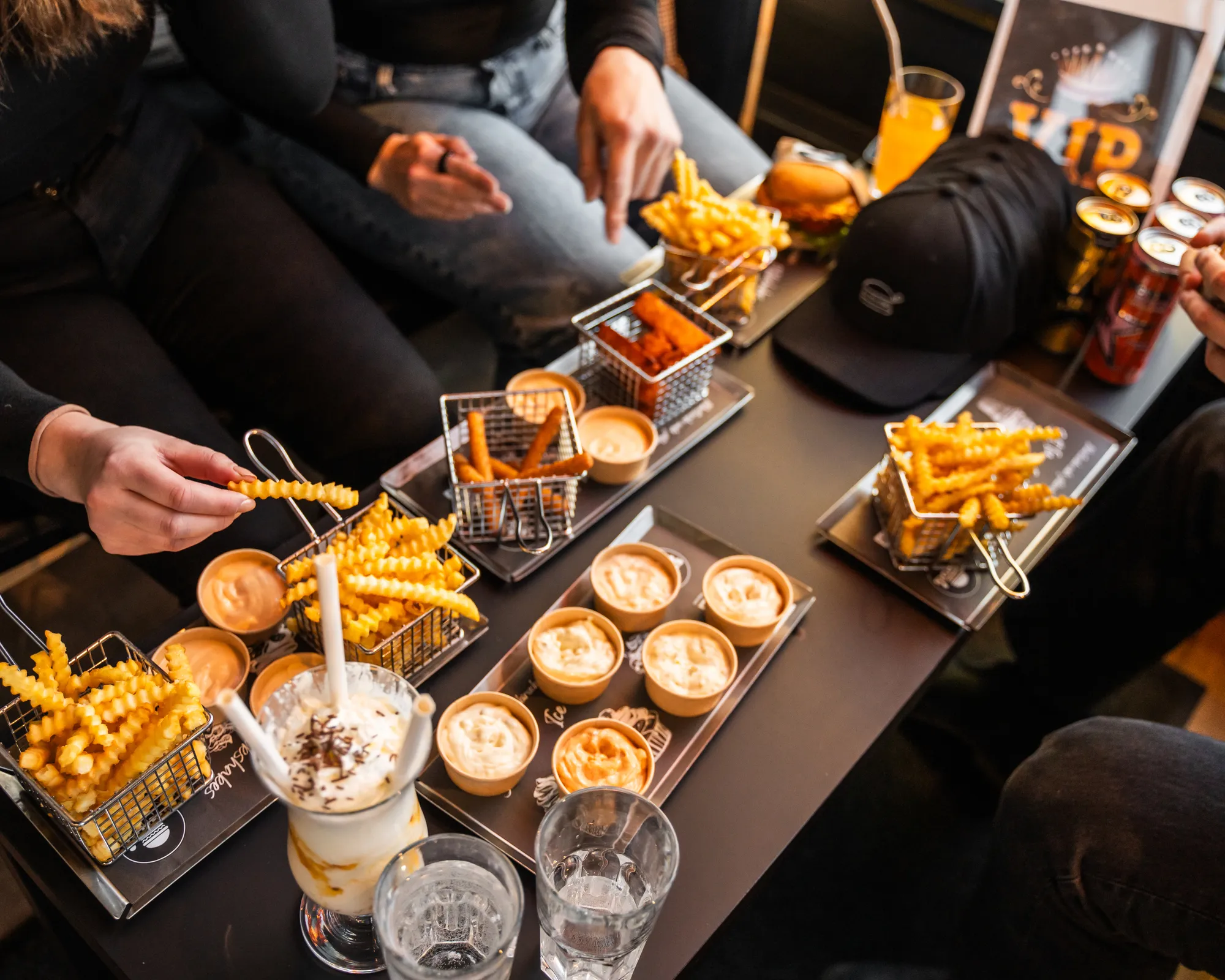 Ryhmä ihmisiä istuu pöydän ympärillä, pöydällä aaltoilevilla reunoilla olevia ranskalaisia, dippikastikkeita, hampurilainen, juomia ja pirtelö.