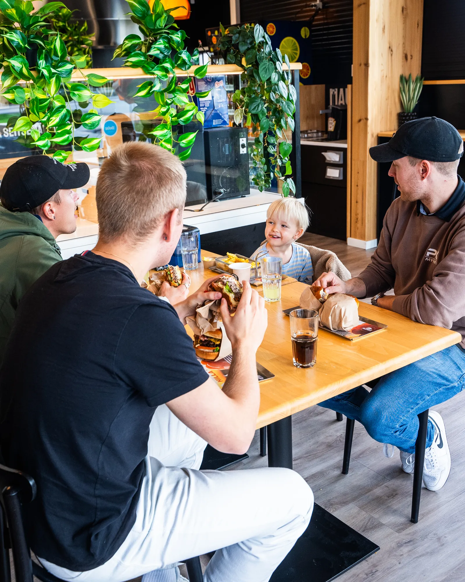 Kolme nuorta miestä ja hymyilevä taapero istuvat puisella pöydällä syöden hampurilaisia ja ranskalaisia rennossa ravintolassa.