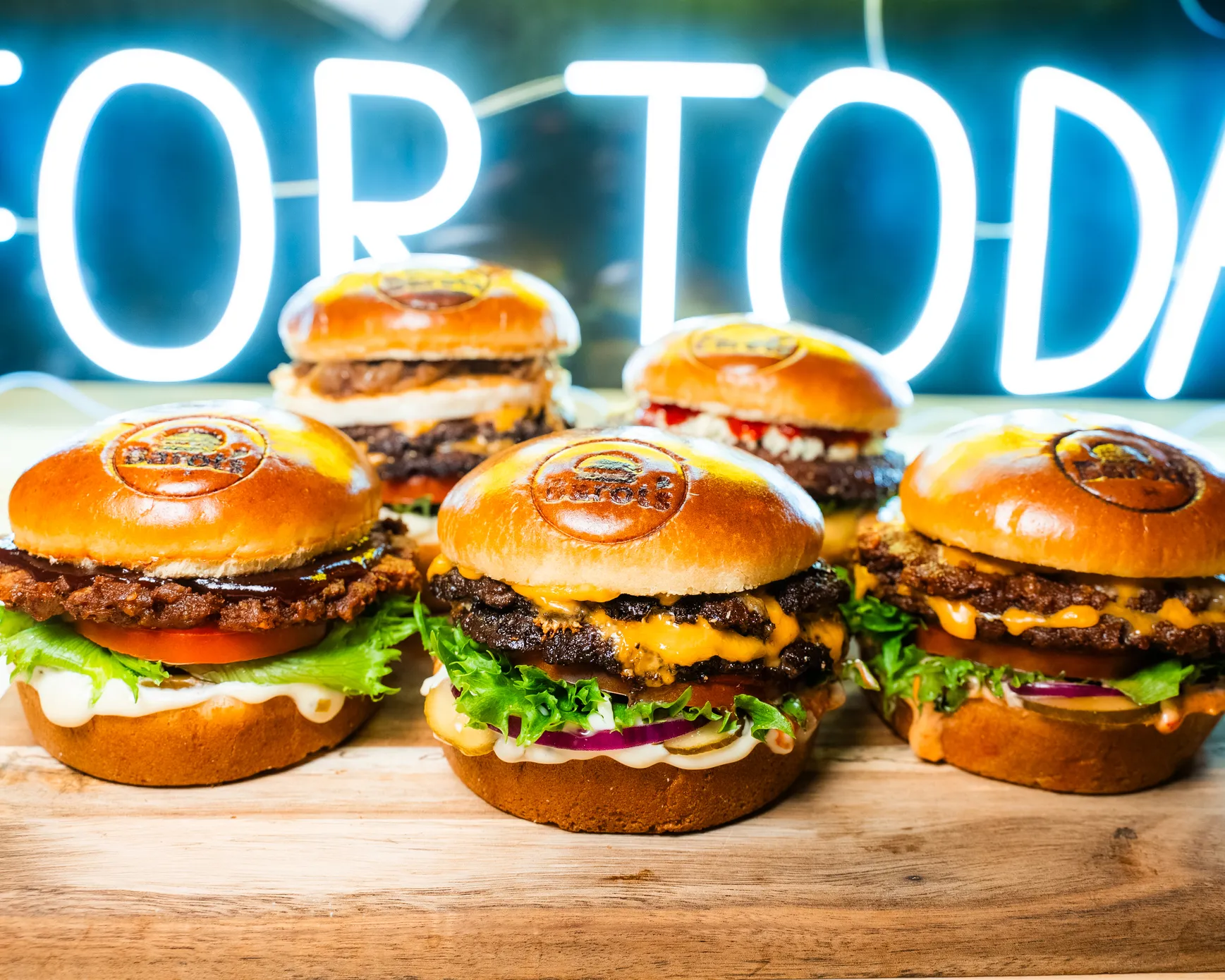 Viisi erilaista hampurilaista brändätyillä sämpylöillä puulautasella, taustalla neonvalo.