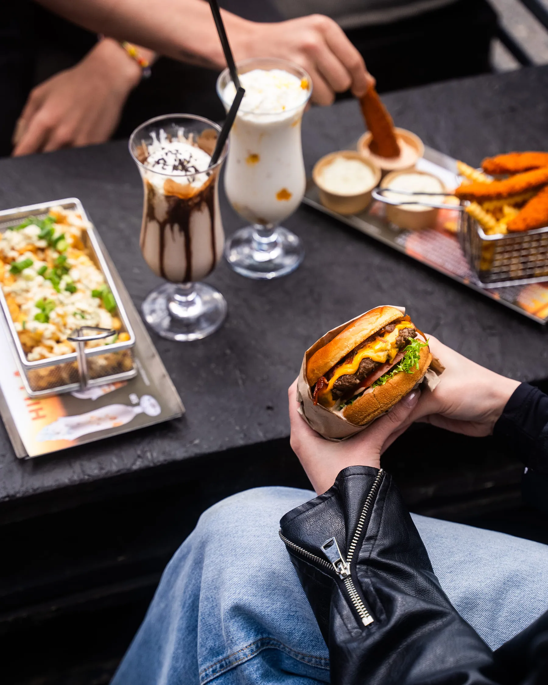 Henkilö pitelee juustohampurilaista, jossa on salaattia, tomaattia ja pekonia; pöydällä mustalla pinnalla on pikaruokaa ja pirtelöitä.