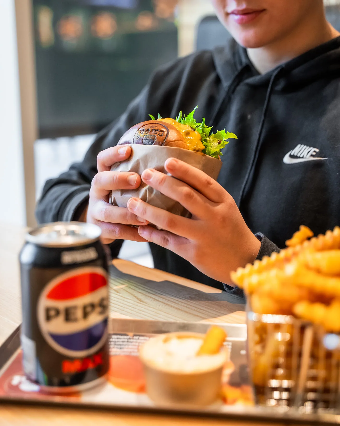 Henkilö mustassa Nike-hupparissa pitelee paperiin käärittyä juustohampurilaista, jossa näkyy salaattia; tarjottimella edessä on tölkki Pepsi Maxia, ranskalaiset metallikorissa ja dippikulho.