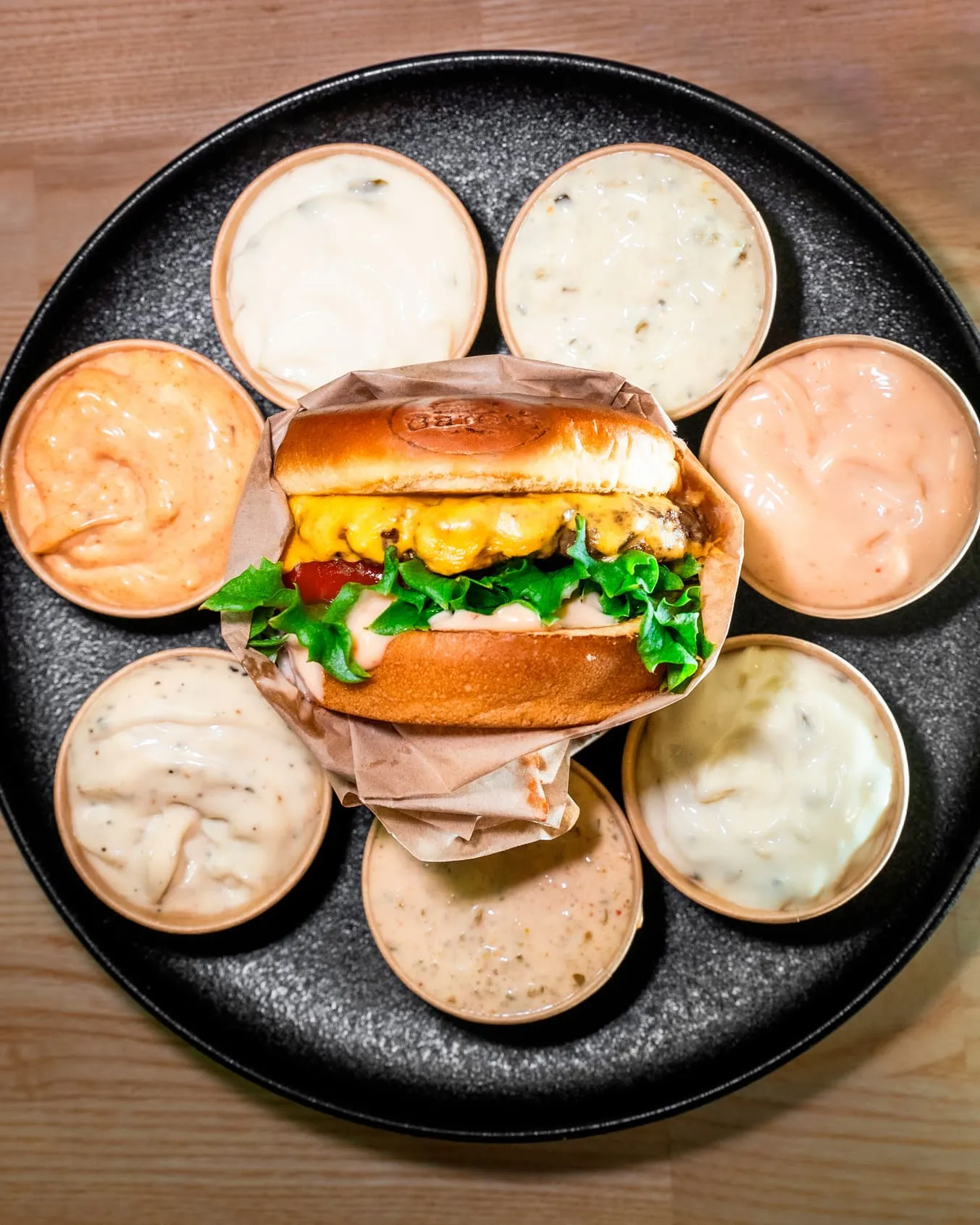 Juustohampurilainen, jossa on salaattia ja tomaattia, mustalla lautasella, ympärillä seitsemän pientä kuppia erilaisia dippikastikkeita.