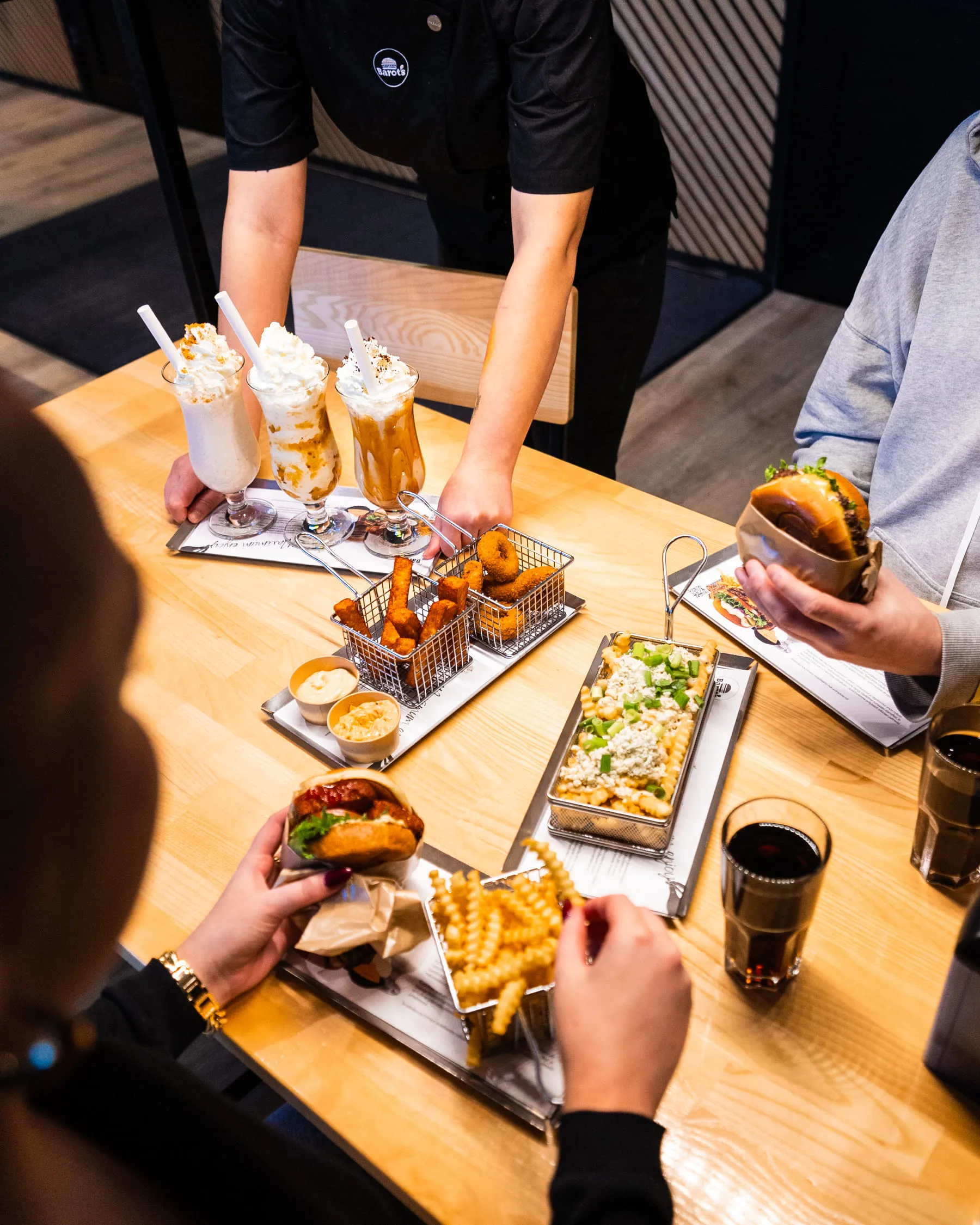 Ihmiset istuvat puisella pöydällä nauttien hampurilaisista, aaltoilevilla reunoilla olevista ranskalaisista, juustolla kuorrutetuista loaded‑ranskalaisista, friteeratuista lisukkeista dippikastikkeiden kanssa ja kermavaahdolla kuorrutetuista pirtelöistä.
