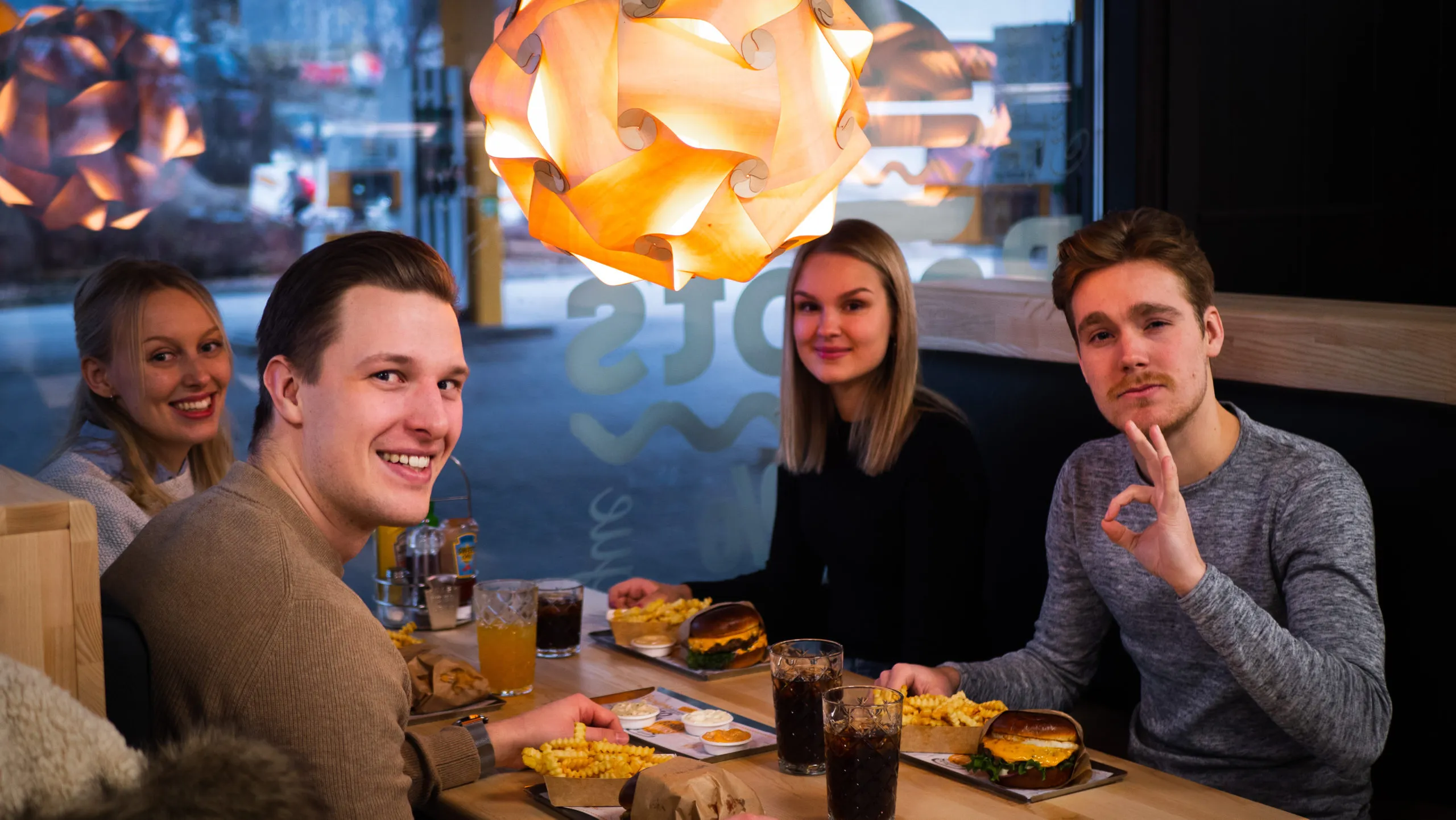 Neljä nuorta aikuista istuu ravintolan nurkkapöydässä nauttien hampurilaisista, aaltoilevilla reunoilla olevista ranskalaisista ja virvoitusjuomista lämpimän värisissä riippuvalaisimissa.