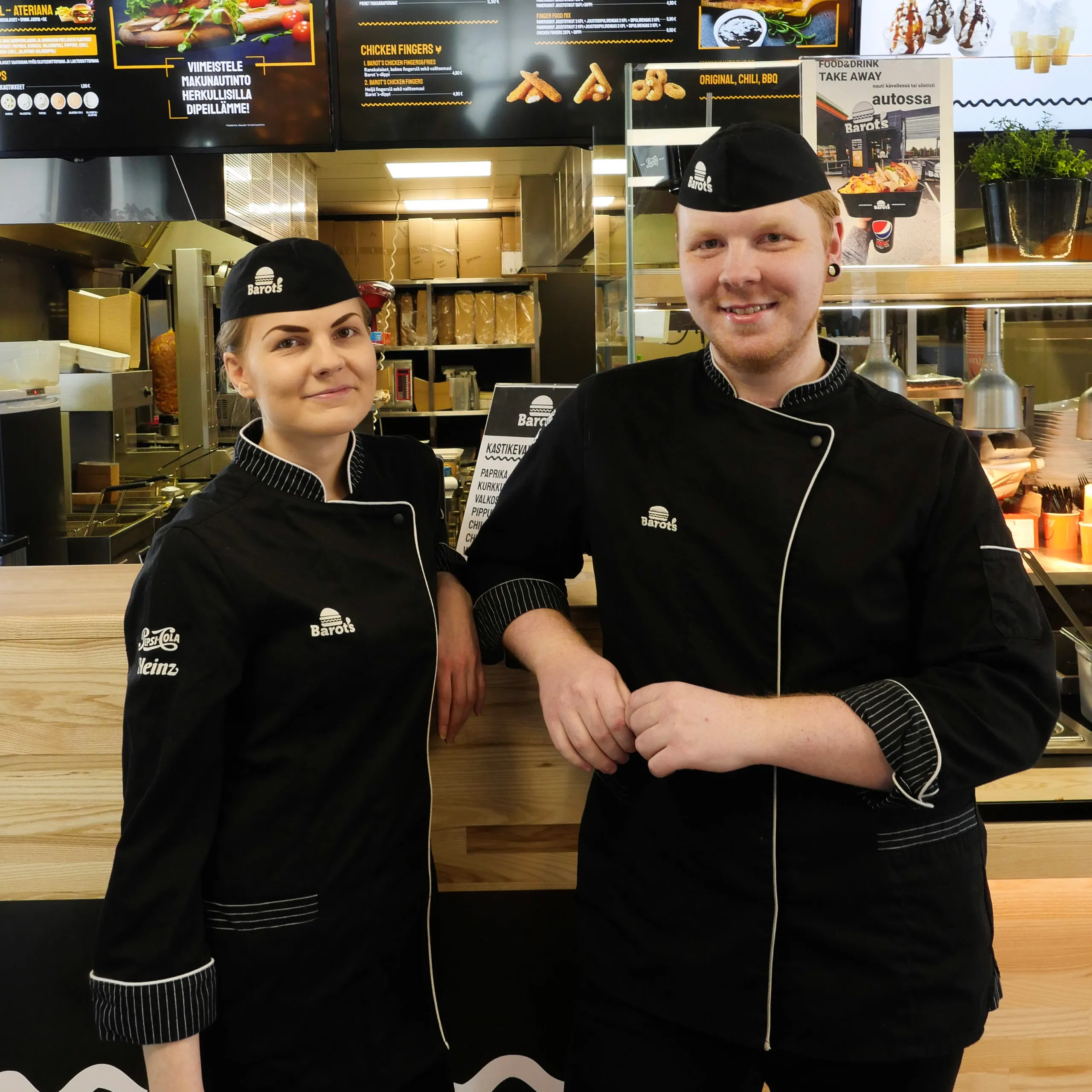 Kaksi hymyilevää pikaruokatyöntekijää, jotka pukeutuneina mustiin Barot’s-uniformuihin ja -lippiksiin, seisoo tiskin edessä ravintolan keittiössä.