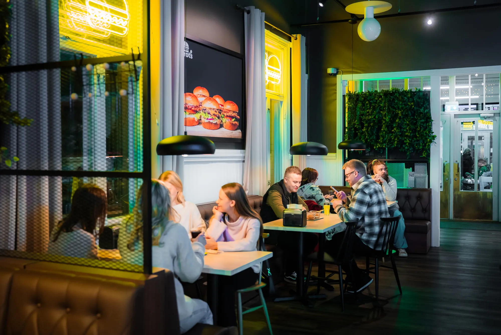 Kolme nuorta miestä ja pieni lapsi istuvat puisella pöydällä syömässä hampurilaisia ja ranskalaisia rennossa ravintolassa, jonka sisustuksessa on riippuvia vihreitä kasveja.