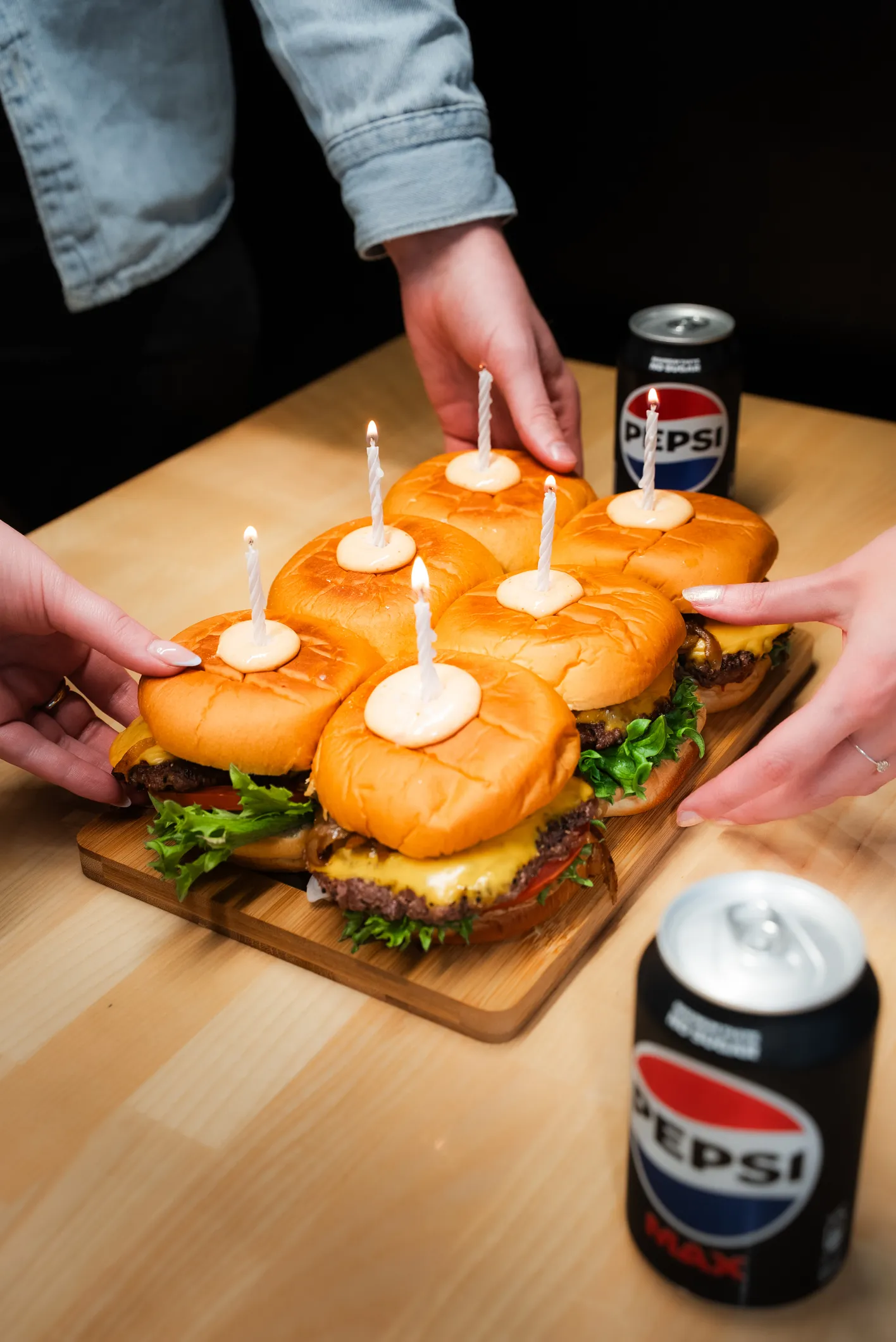 Viisi juustohampurilaista, joiden päällä palaa syntymäpäiväkakkuun tarkoitetut kynttilät, puulautasella; ympärillä näkyy käsiä ja kaksi tölkkiä Pepsi Maxia vaalealla puupöydällä.