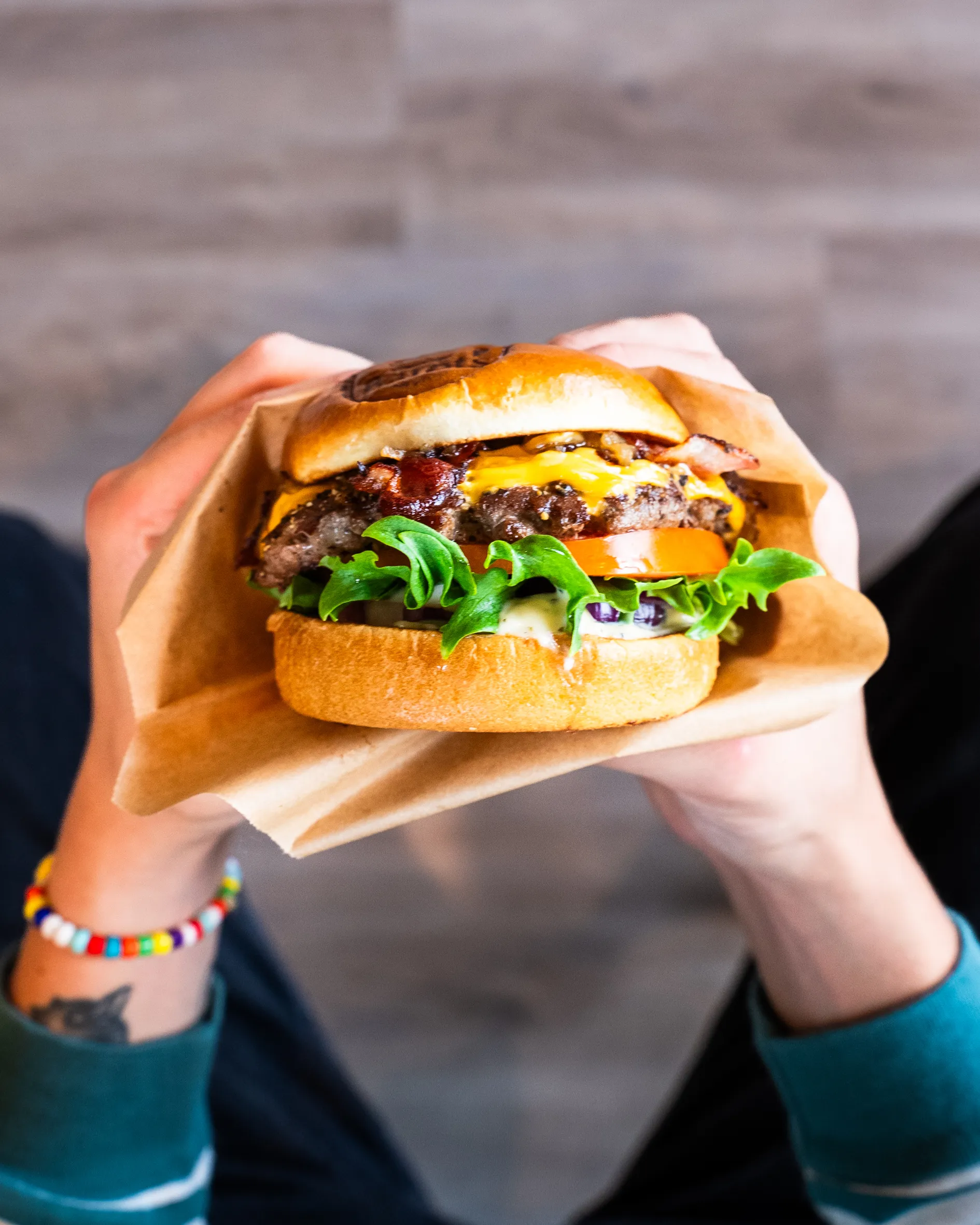 Henkilö pitelee juustohampurilaista, jossa on pekonia, salaattia, tomaattia ja kastiketta, käärittynä ruskeaan paperiin.