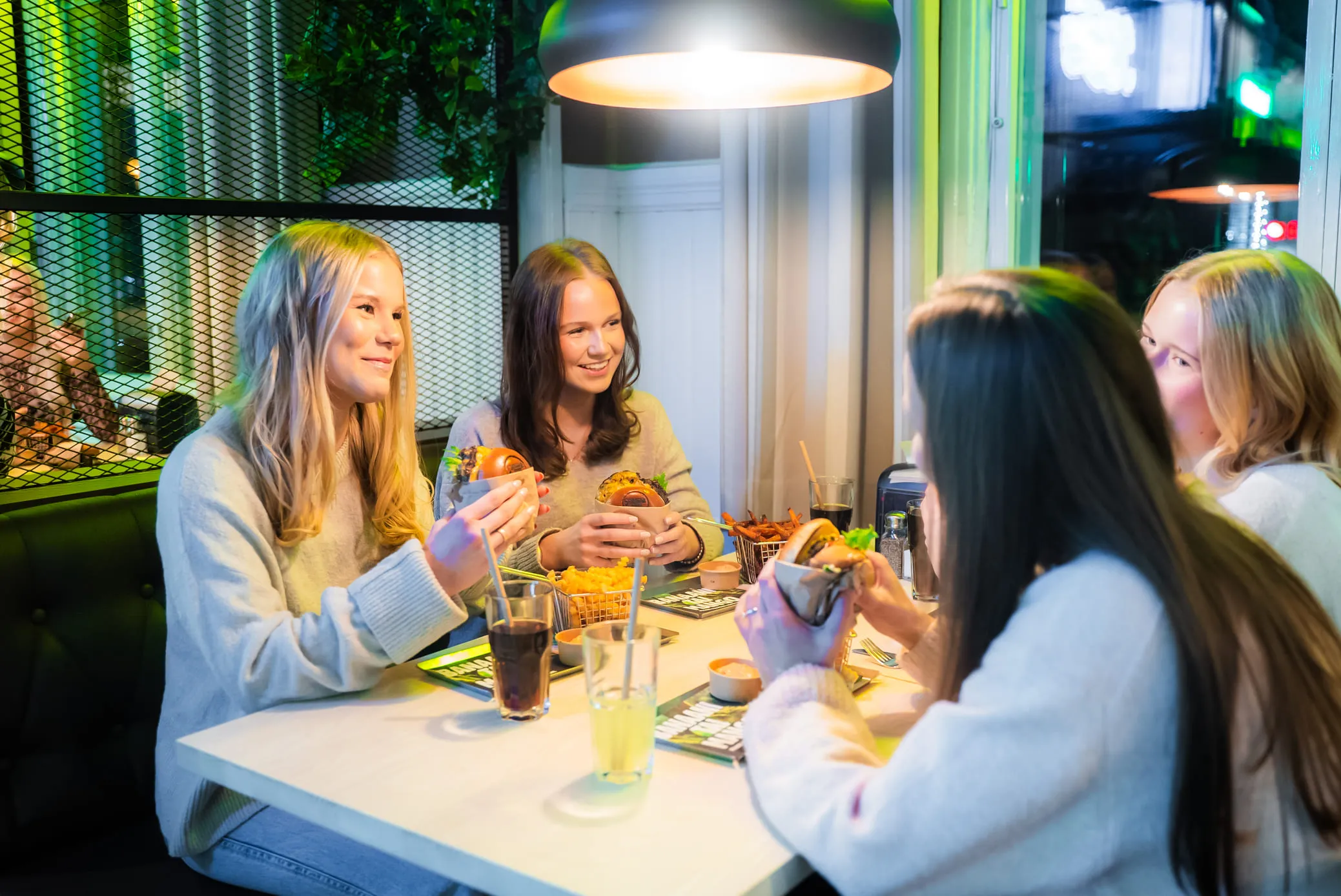 Neljän naisen ryhmä istuu pöydän ympärillä ravintolassa, pitäen hampurilaisia ja hymyillen; pöydällä on juomia ja ranskalaisia, ja yläpuolella roikkuu kattovalaisin.