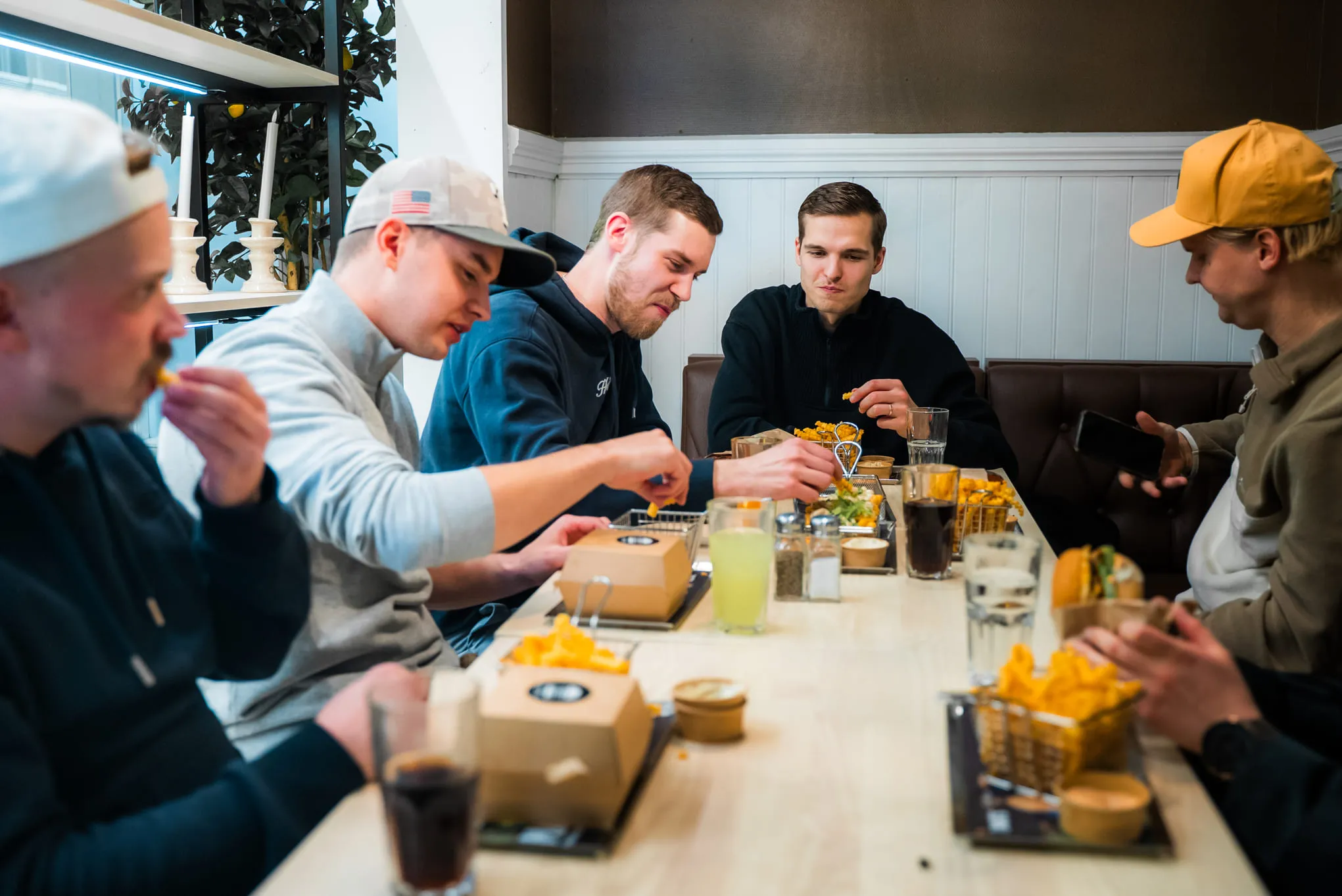 Kuusi nuorta miestä istuu pöydän ympärillä syöden rentoa pikaruokaa, kuten hampurilaisia, ranskalaisia ja juomia, viihtyisässä ravintolaympäristössä.