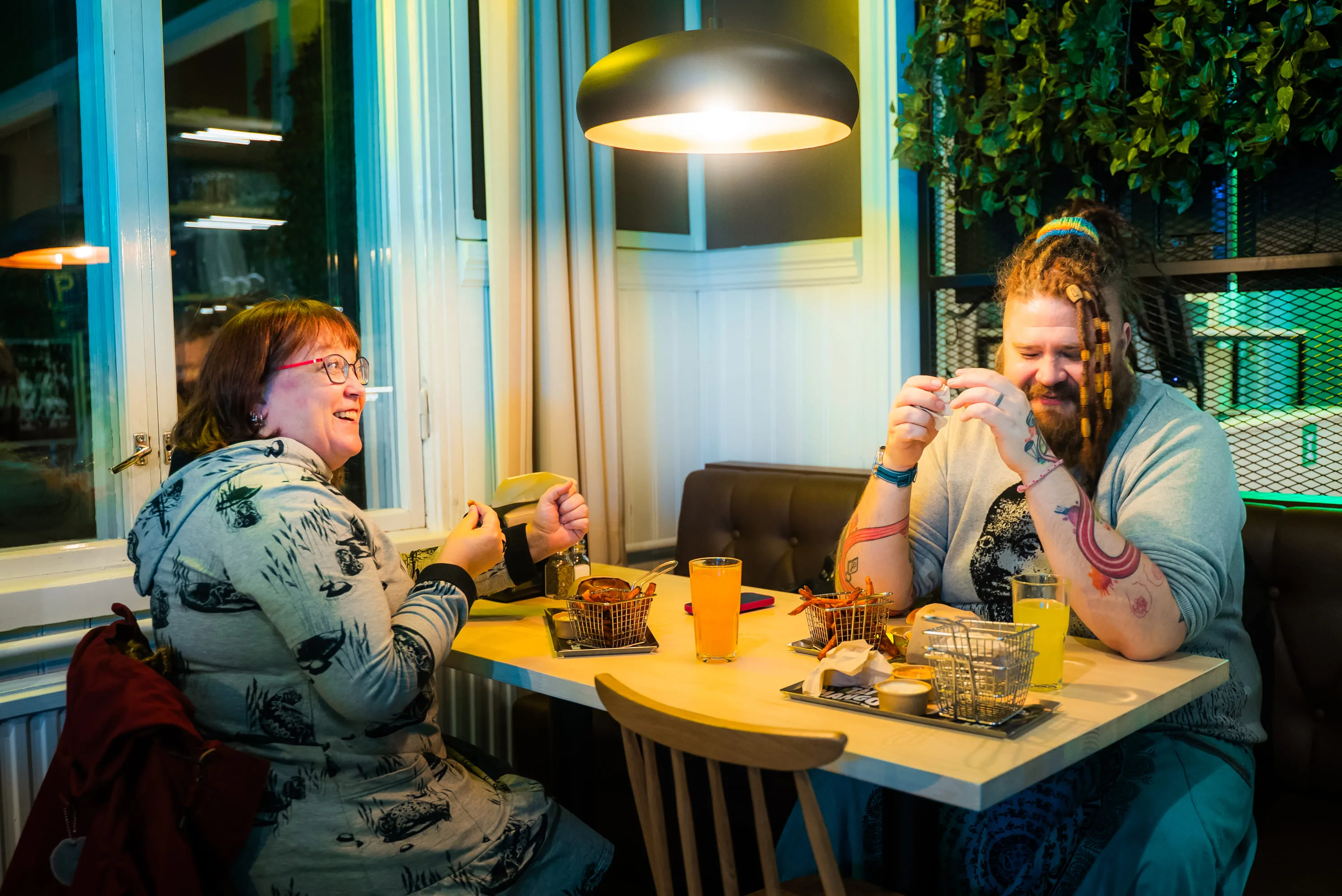 Kaksi ihmistä istuu ravintolapöydässä, pöydällä ruokakorit ja juomia, nauttien vilkkaasta keskustelusta kattovalaisimen alla.