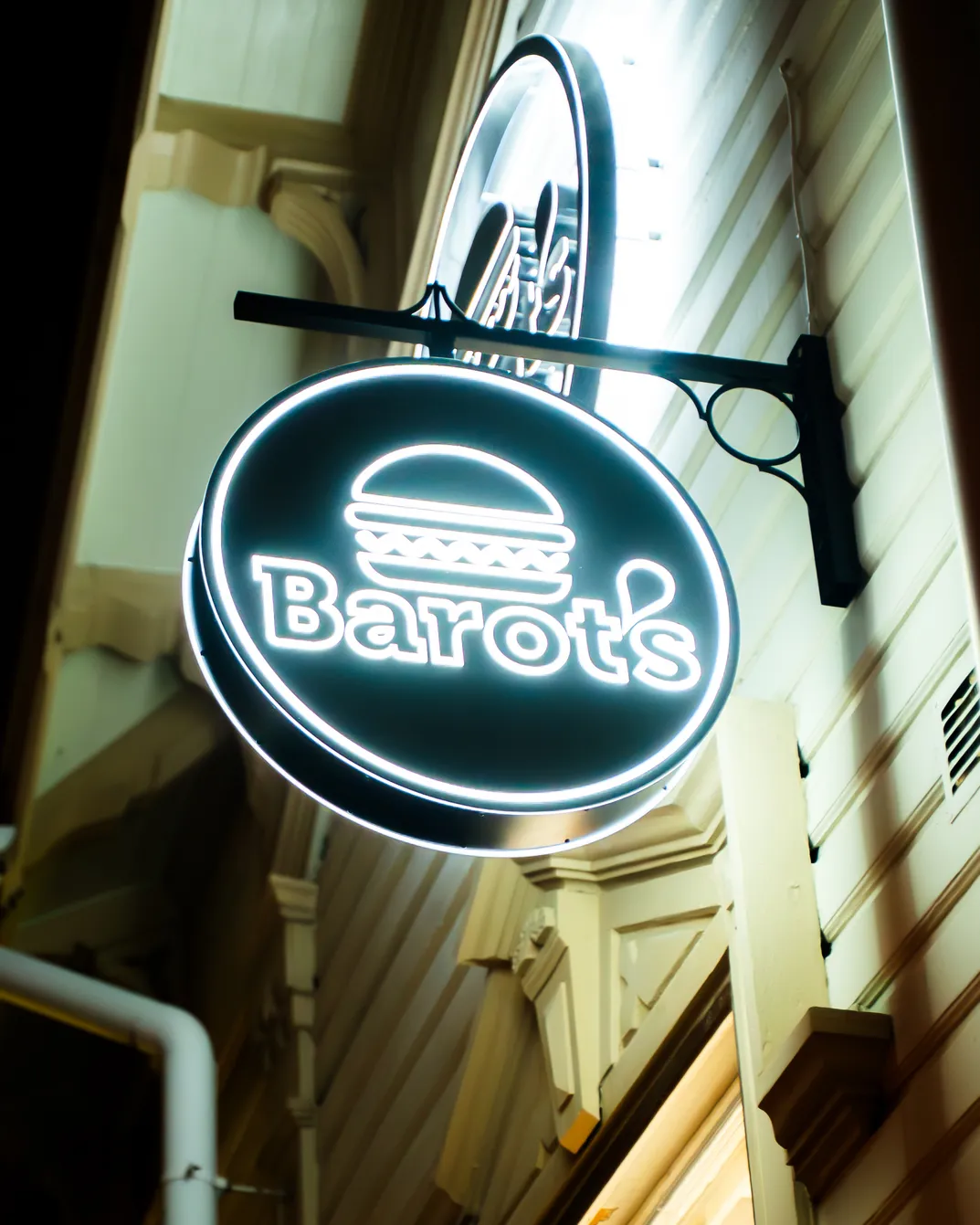 Valaistu pyöreä kyltti, jossa on hampurilaisikoni ja sana "Barot's", roikkumassa rakennuksen ulkopuolella yöllä.