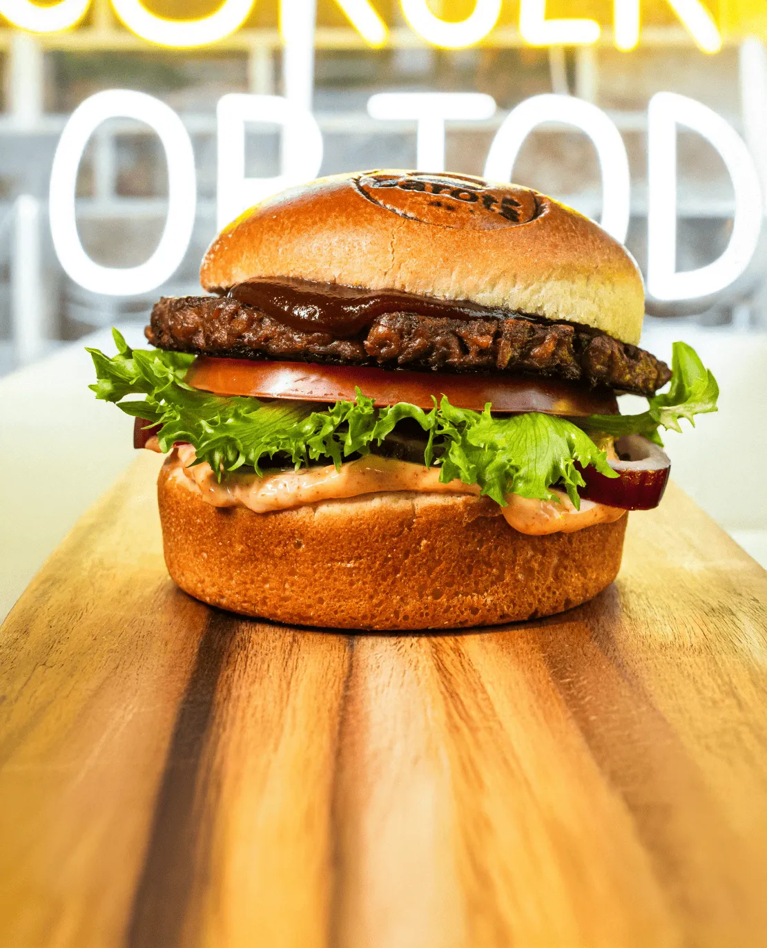 Kasvishampurilainen, jossa on salaattia, tomaattia, sipulia, kastiketta ja BBQ-kastiketta paahdetussa sämpylässä, asetettuna puiselle alustalle.