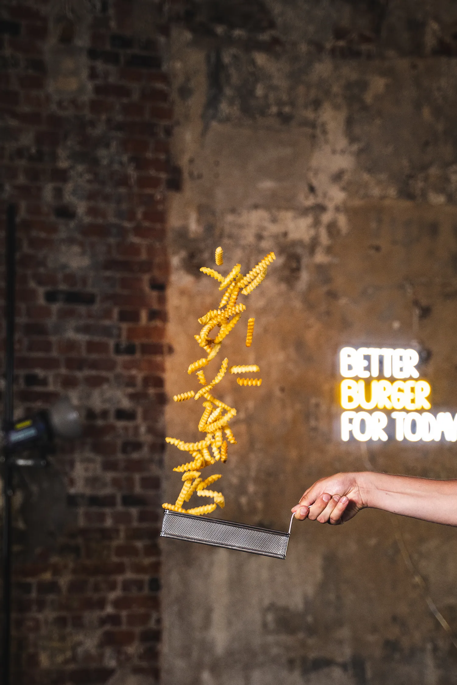 Lähikuva juustohampurilaisesta, jossa on salaattia, tomaattia, sipulia ja barbecue-kastiketta, tarjoiltuna puisella pöydällä ja valaistuna neonilla olevasta burger-kyltistä.