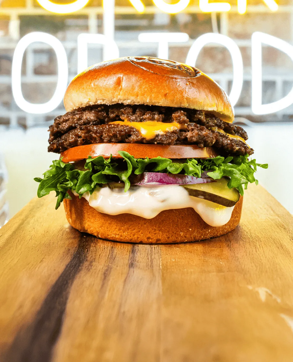 Tuplajuustohampurilainen, jossa on salaattia, tomaattia, sipulia, suolakurkkua ja kastiketta puisella leikkuulaudalla.