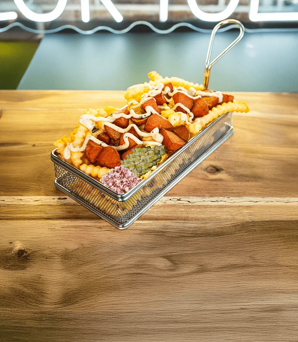 Pieniä paistettuja perunoita ja makkarapaloja kastikkeella koristeltuna, tarjoiltuna metallikorissa silputtujen suolakurkkujen ja sipulin kanssa.