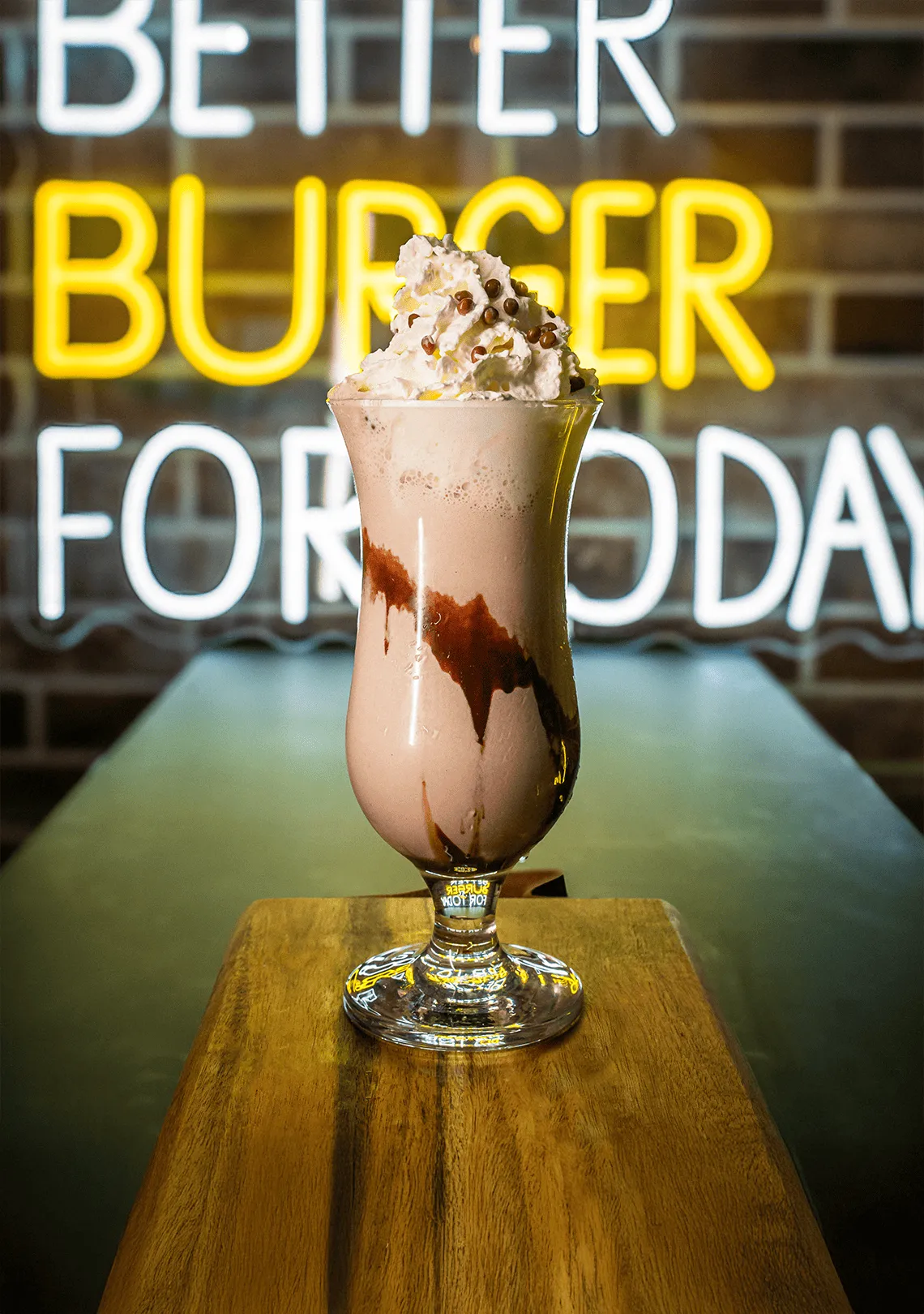 Suklaamaitoshake korkeassa lasissa, päällä kermavaahtoa ja suklaahippuja, asetettuna puiselle laudalle.