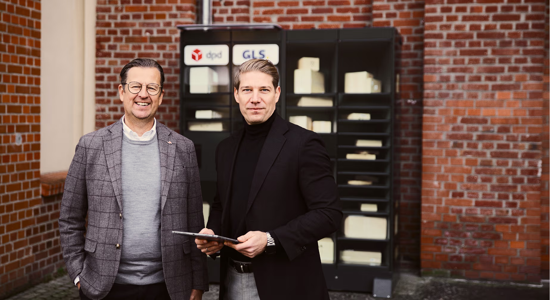 Zwei Männer vor einer inboxx Paketstation mit den Logos von dpd und GLS, einer hält ein Tablet.