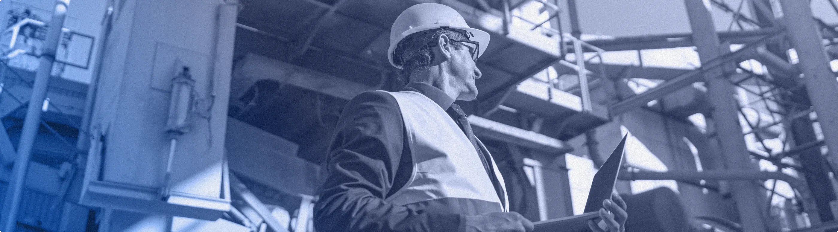 Engineer in a hard hat and safety vest holding a laptop inside an industrial facility.