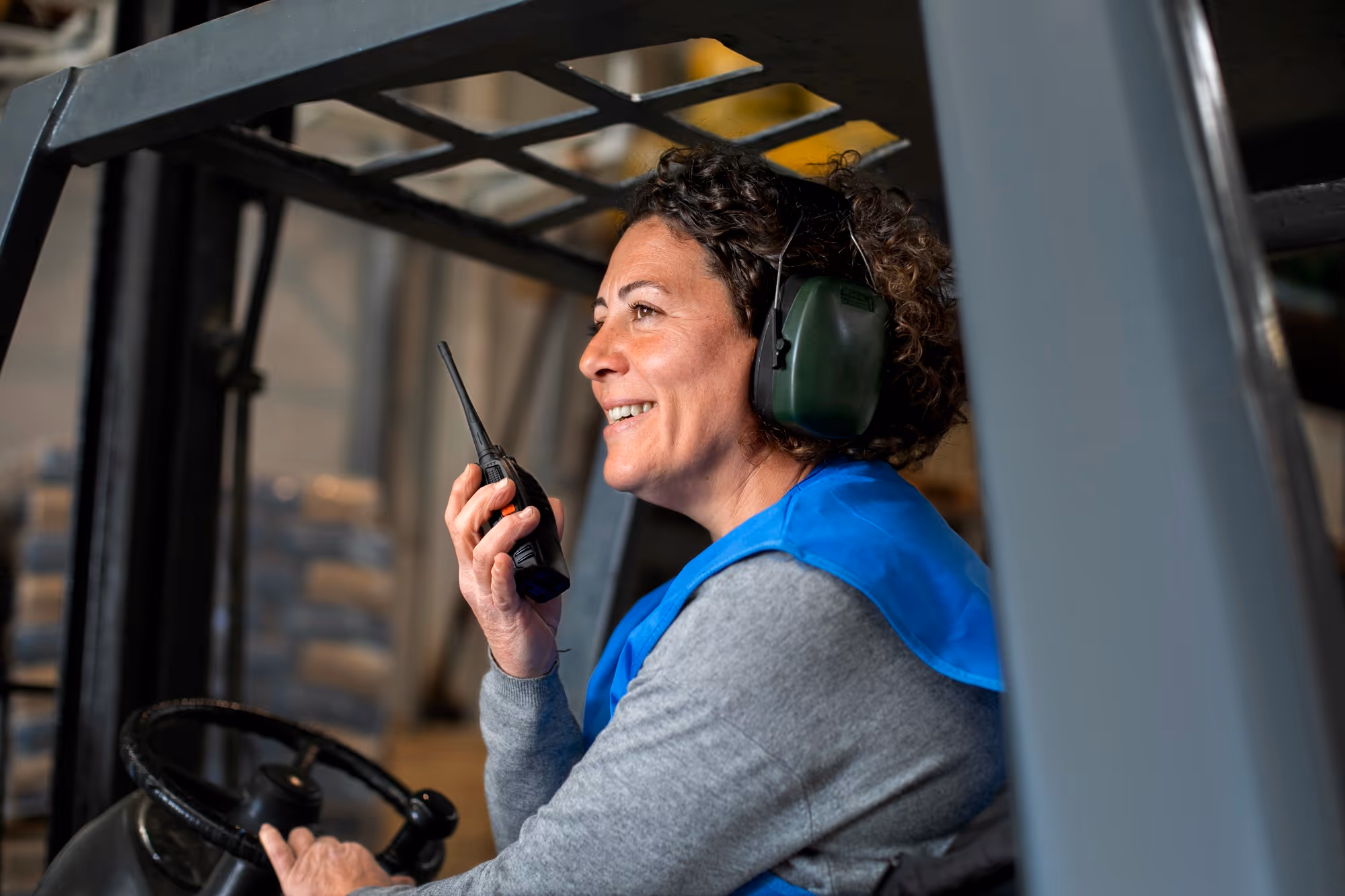 Woman wearing hearing protectors and blue vest using a walkie-talkie while operating a forklift inside a warehouse.