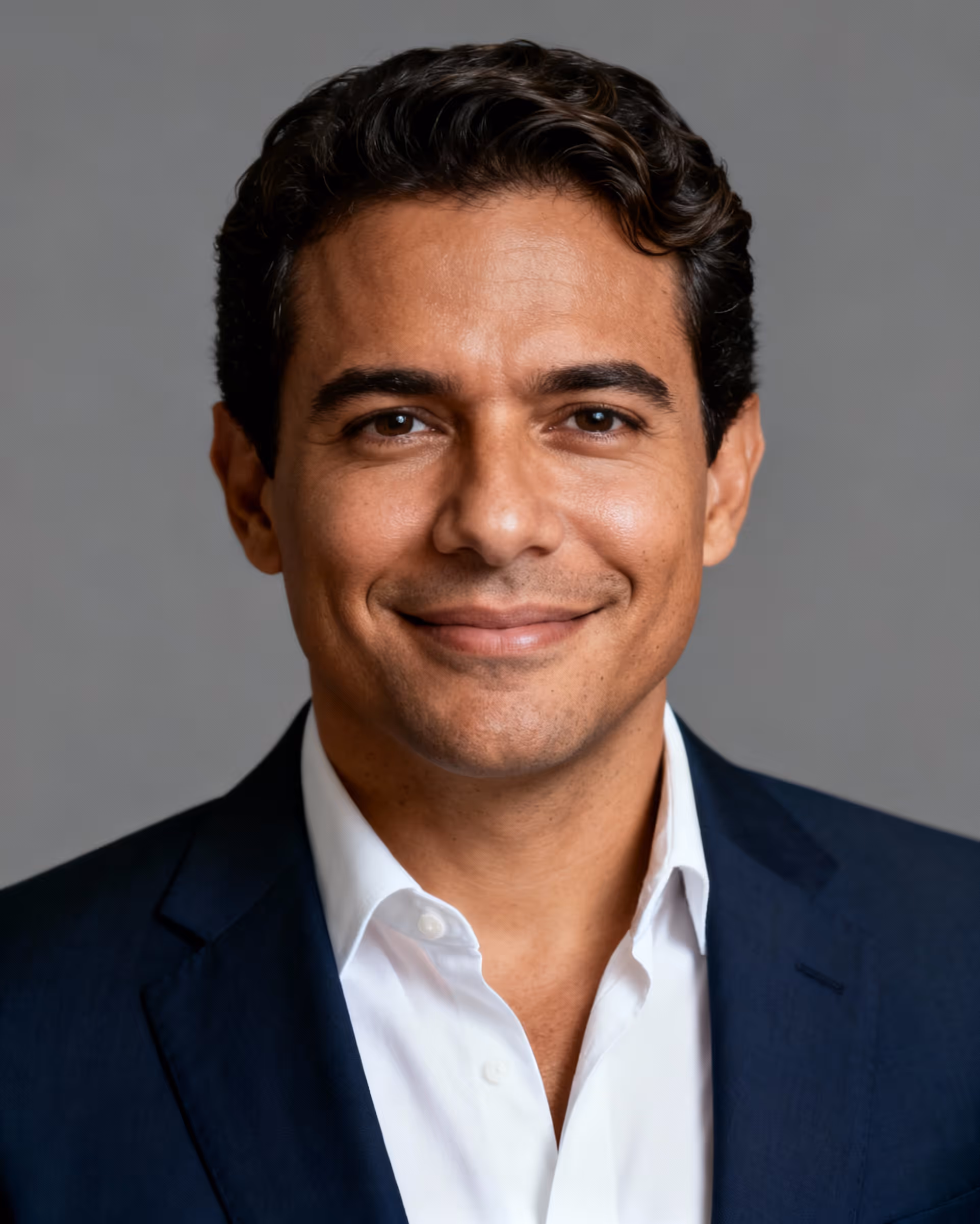 Smiling man with dark curly hair wearing a navy blazer and white shirt against a gray background.