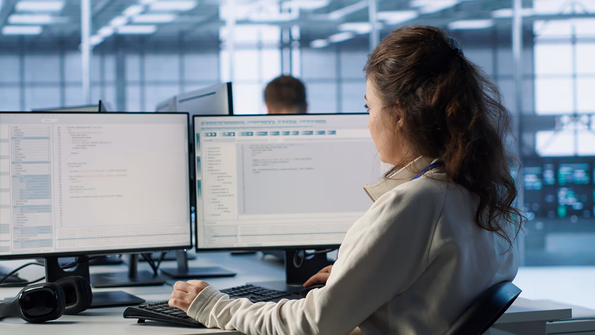 Woman working on dual monitors displaying code in a modern office environment.