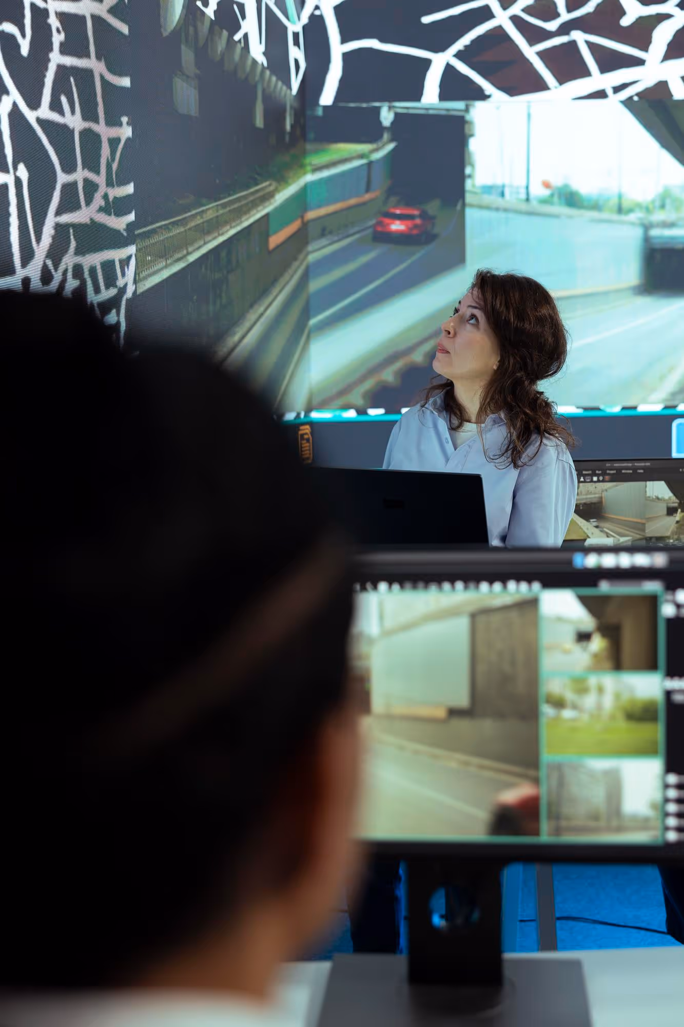 Woman in white shirt looking at a large screen displaying a road and a red car, with a blurred person and computer monitor in the foreground.