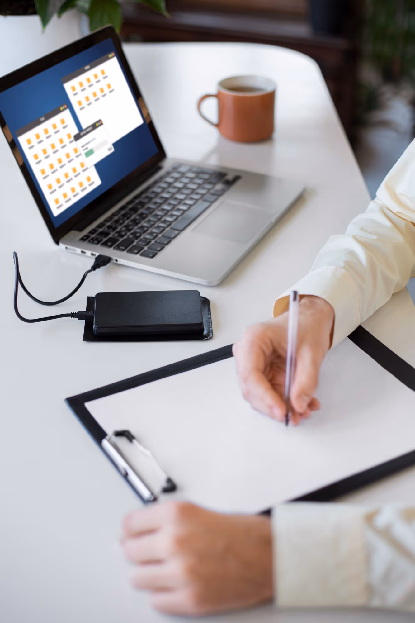Person writing on blank paper on clipboard at a table with a laptop showing file folders and an external hard drive connected.