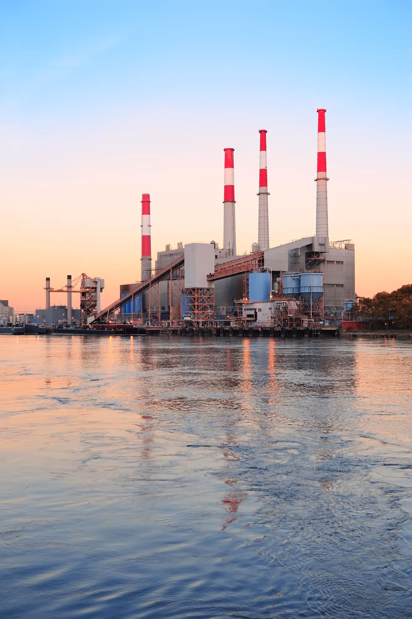 Power plant with four tall smokestacks reflecting on calm water at sunset.