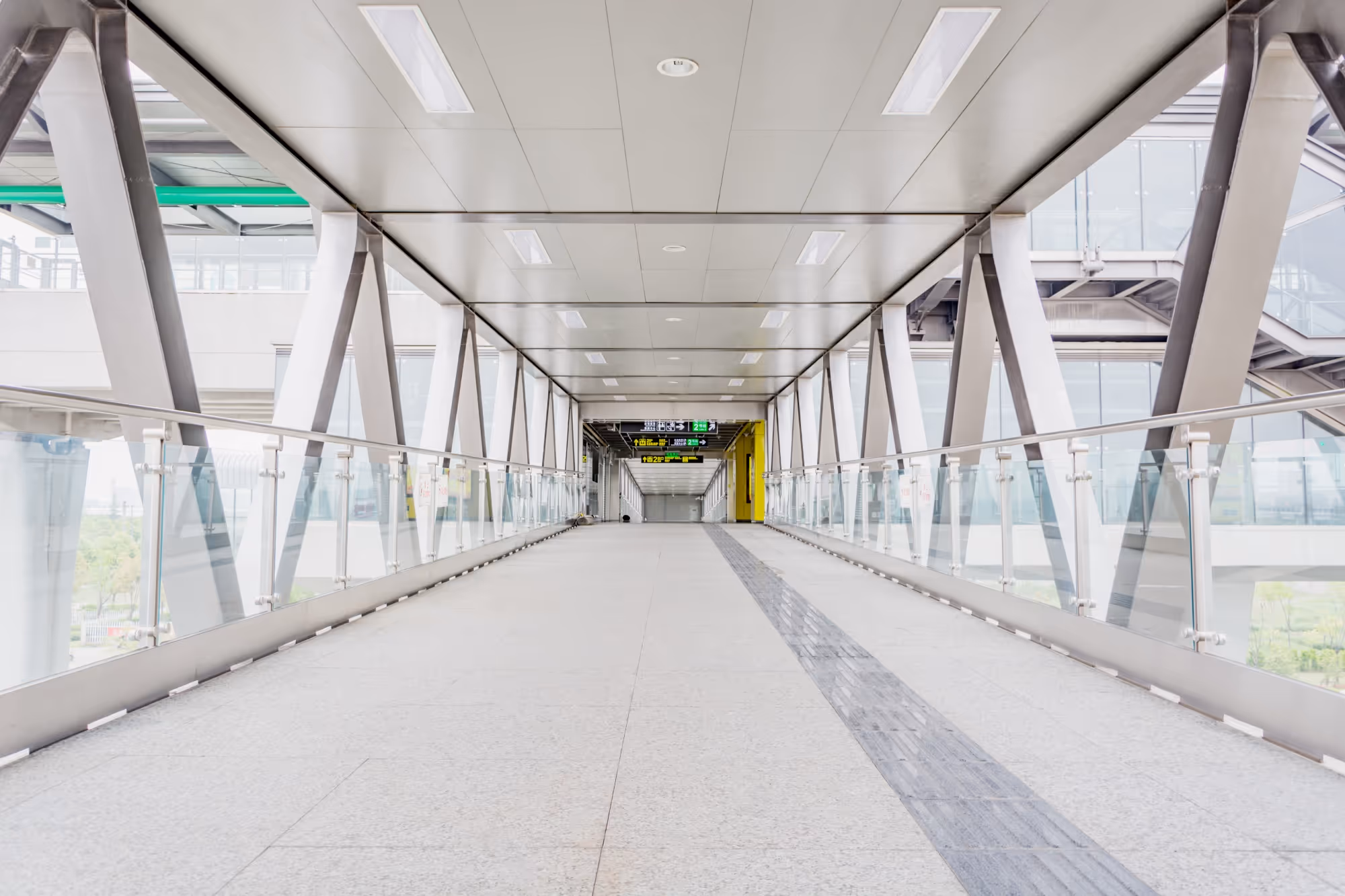 Empty modern pedestrian bridge with glass railings and overhead signs in a commercial or transit building.